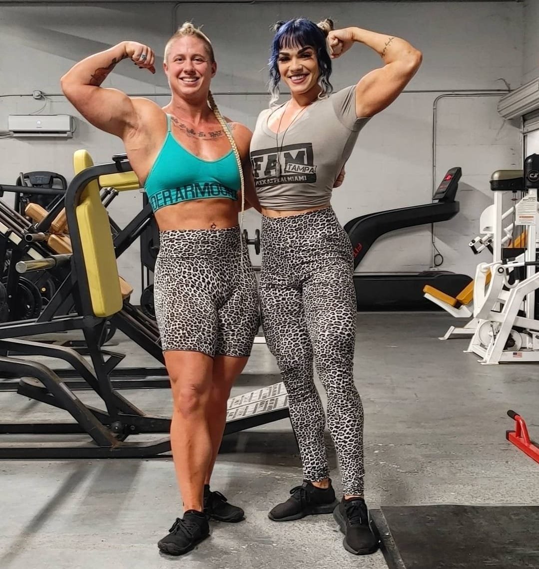 Two women with muscular physiques flexing their arms and smiling at the gym, wearing leopard print leggings and workout tops.