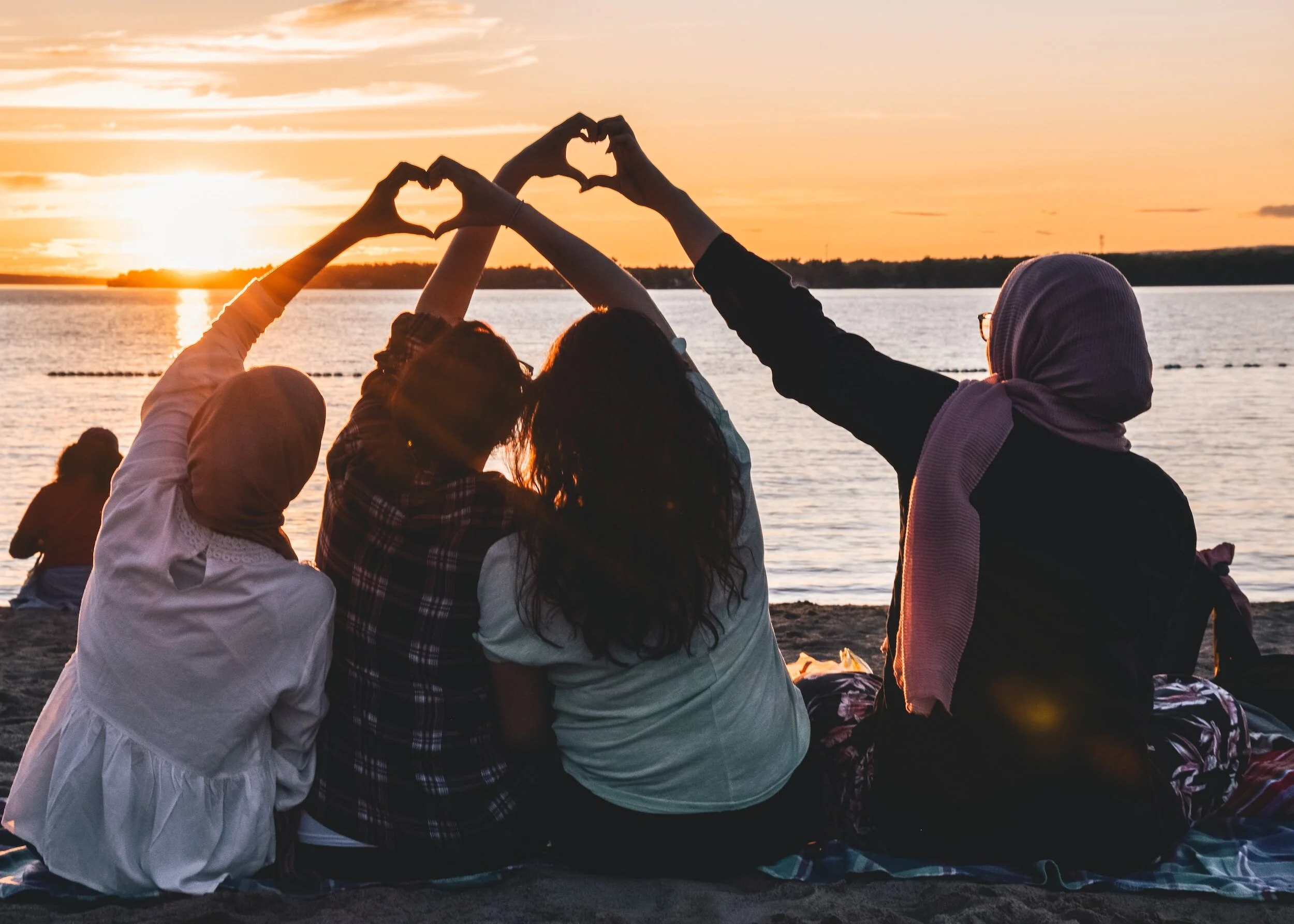 Group of women sitting on a beach at sunset, forming heart shapes with their hands over the water.