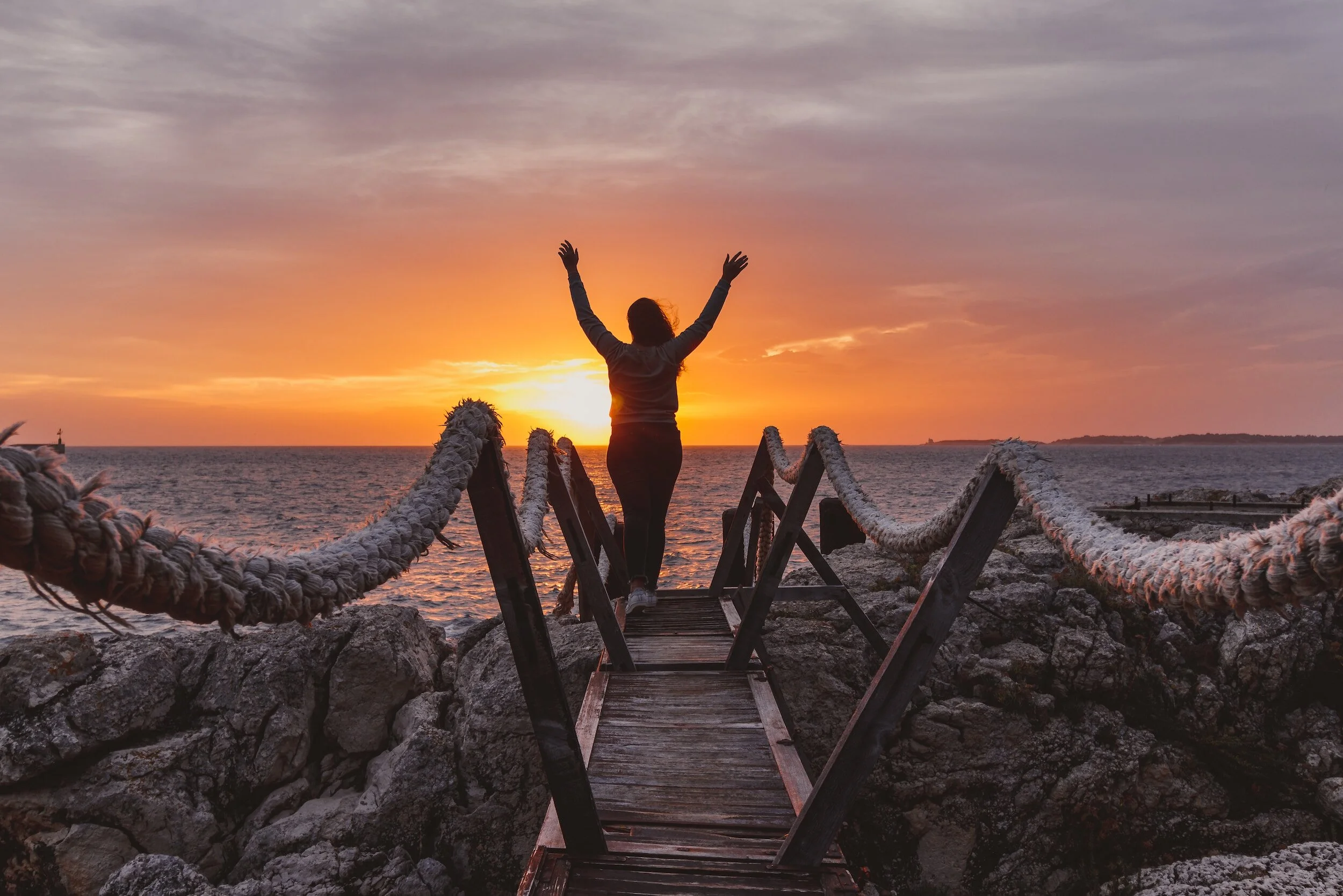 A woman standing on a wooden bridge with arms raised, facing a sunset over the ocean, with rocky terrain and a rope railing.