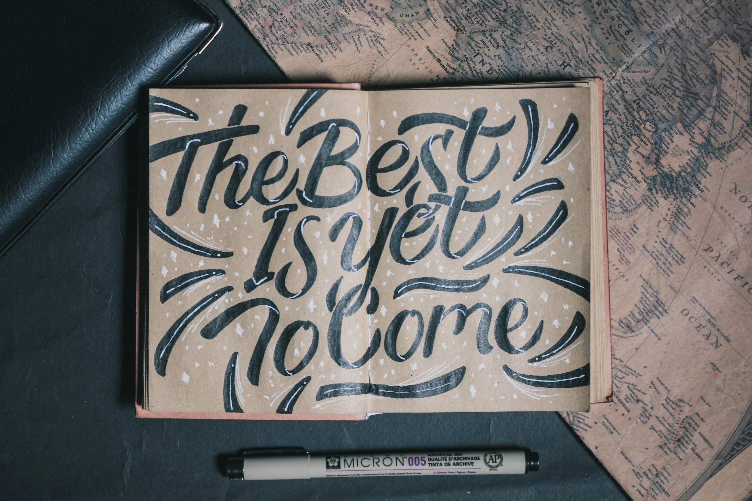 Open notebook with hand-lettered quote, 'The best is yet to come,' written in black and white on brown paper. Surrounded by a black wallet, vintage maps, and a black and white marker, on a dark surface.