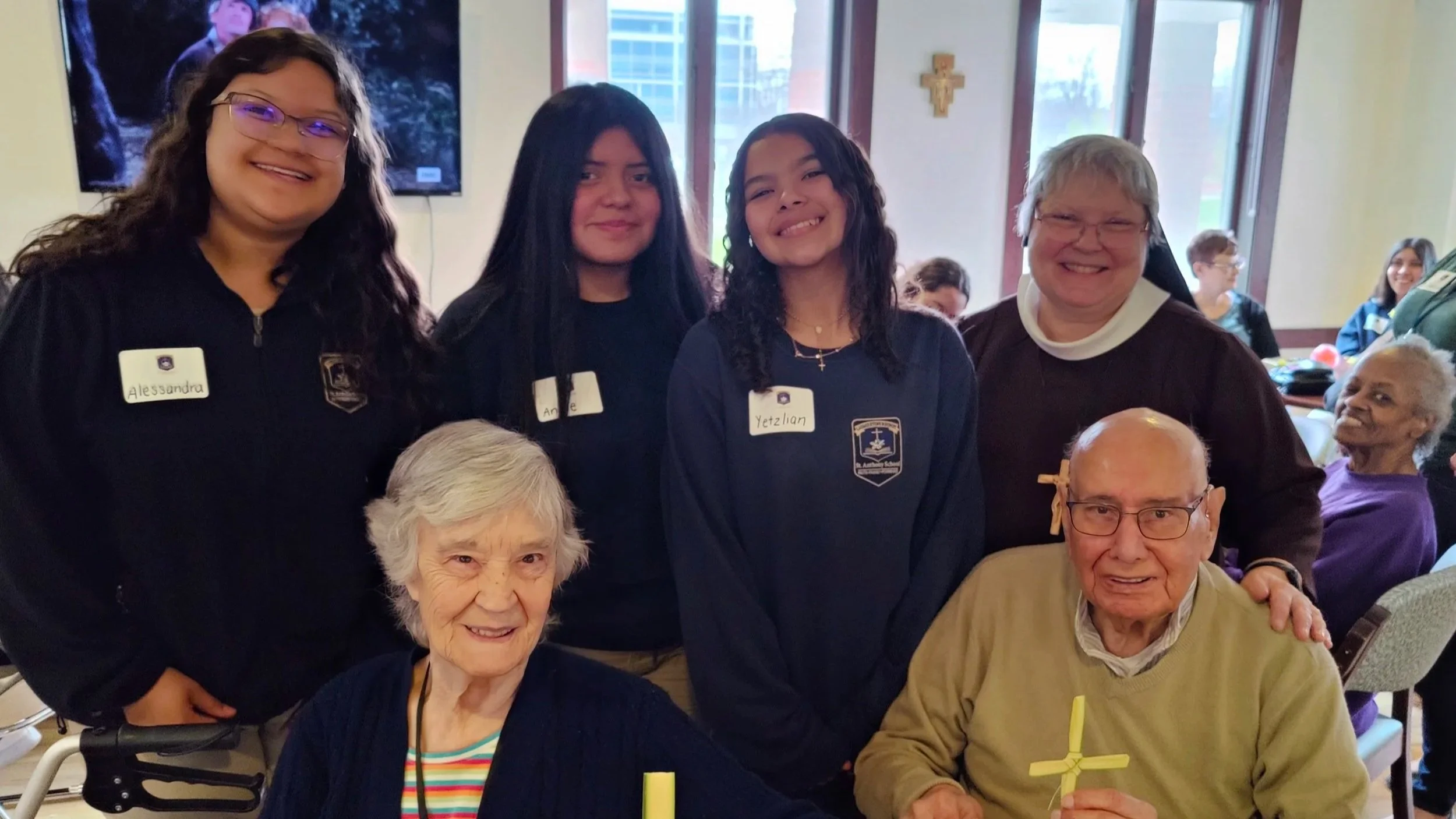 Group of six people, including elderly and young women, gathered indoors. Some are wearing navy blue shirts with name tags, and two men are holding yellow crosses.