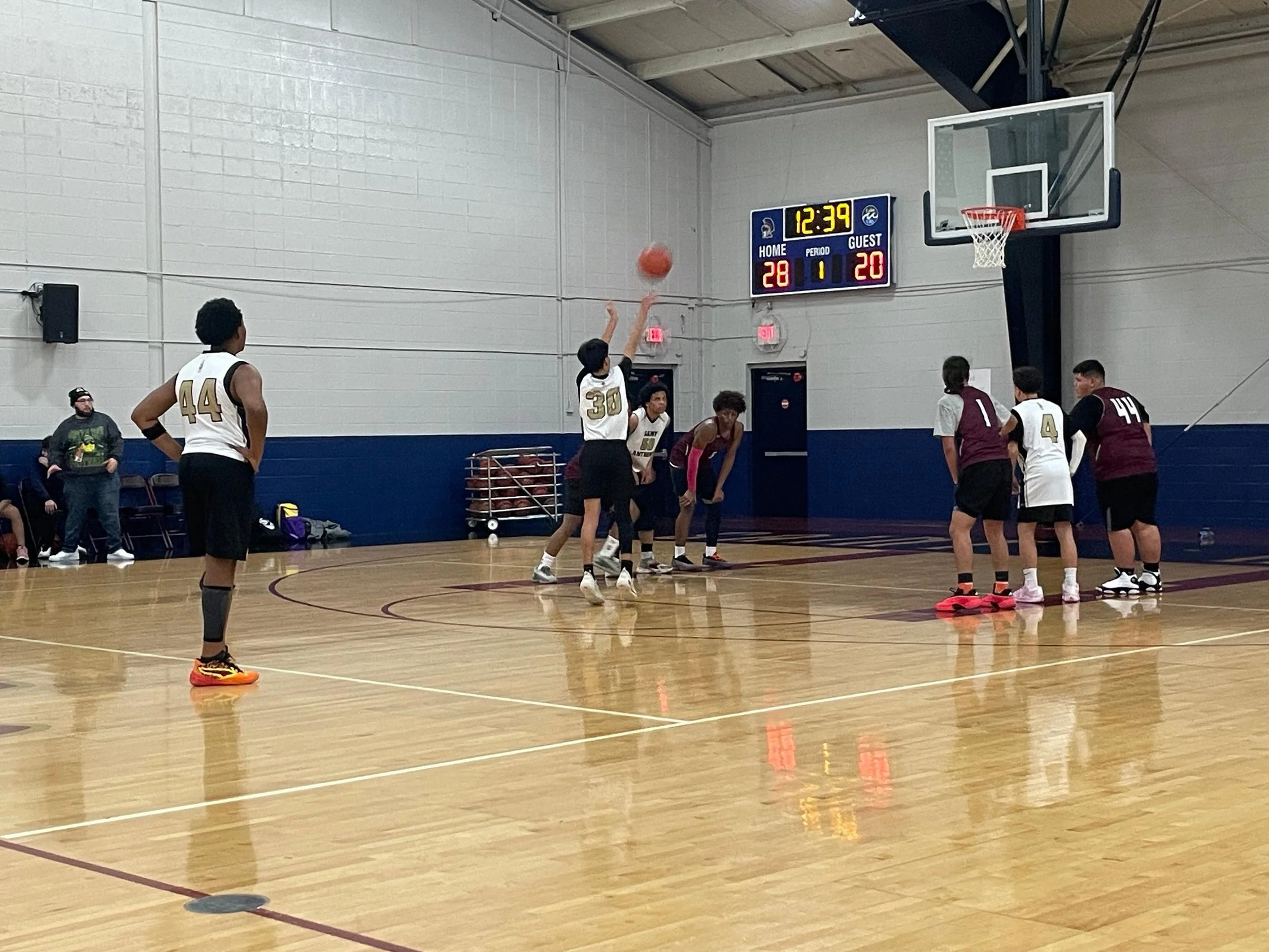 A basketball game in progress with players on the court. One player is shooting the ball, others are defending or waiting. The scoreboard shows 12:39 remaining, 28-20 score, and game period 1.