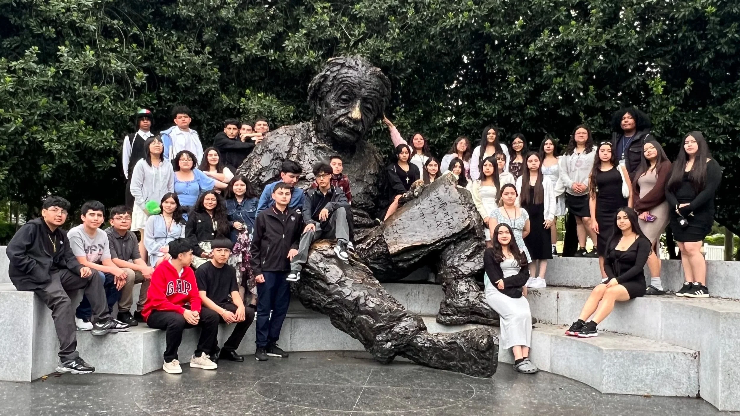 A large group of students surrounding a bronze statue of a man reading a book, seated on a large rock, outdoors with trees in the background.