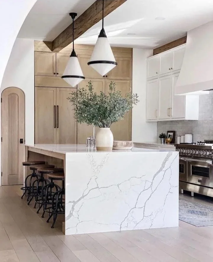 Modern kitchen with a white marble island, light wood cabinets, white upper cabinets, and stainless steel stove. Decorative green plant in a white vase on the island, and two pendant lights hanging above.