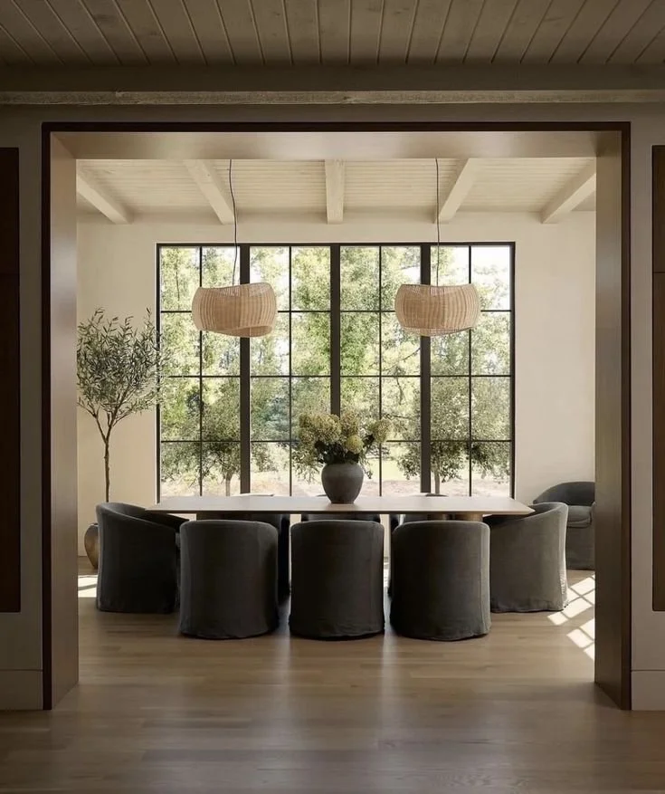 Modern dining room with a large wooden table, eight dark upholstered chairs, and a potted plant on the left side. Large windows in the background allow natural light, with greenery outside. Two hanging pendant lights are above the table.