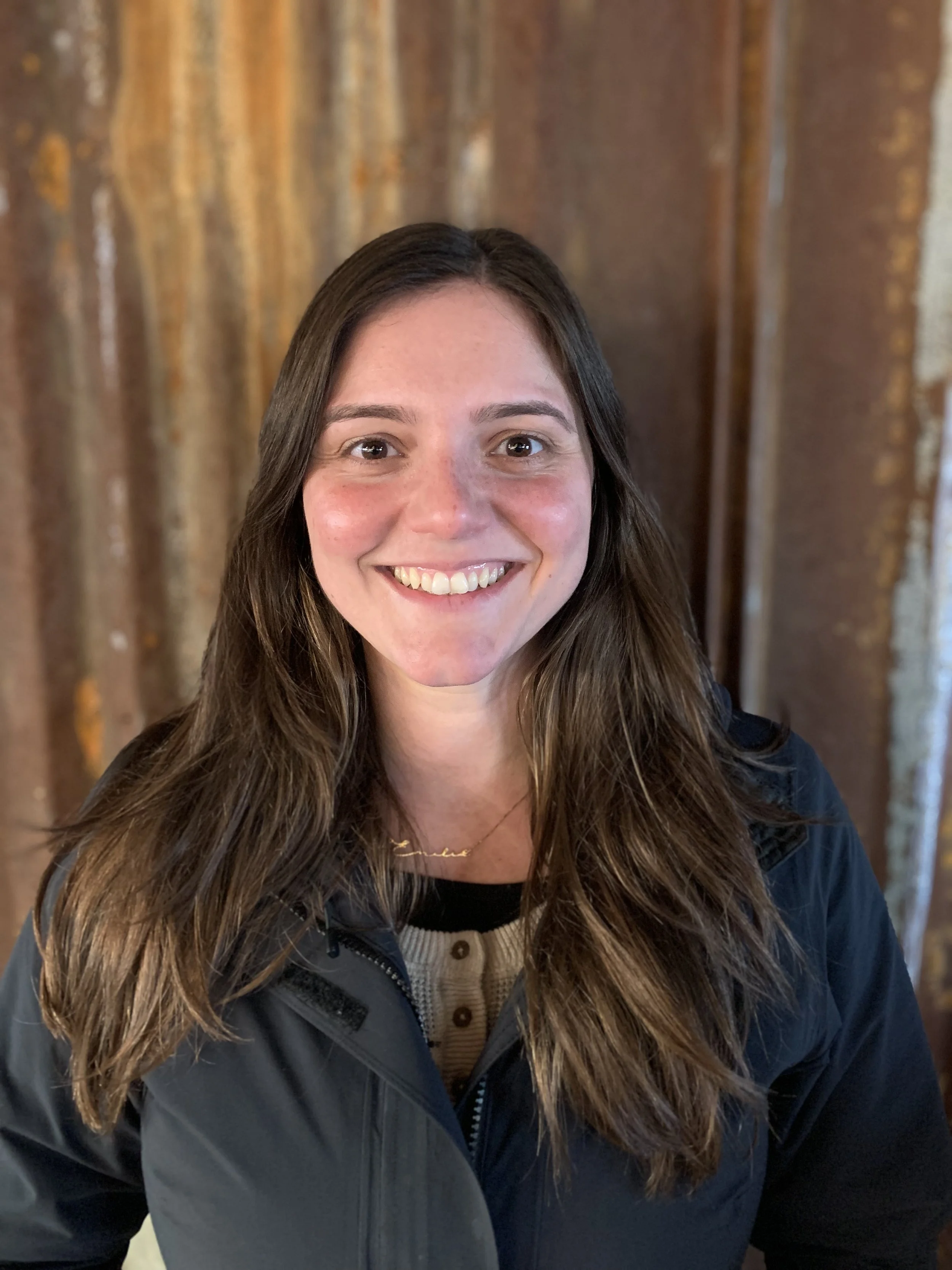 Smiling woman with long brown hair wearing a dark jacket, posing in front of a rustic metal background.