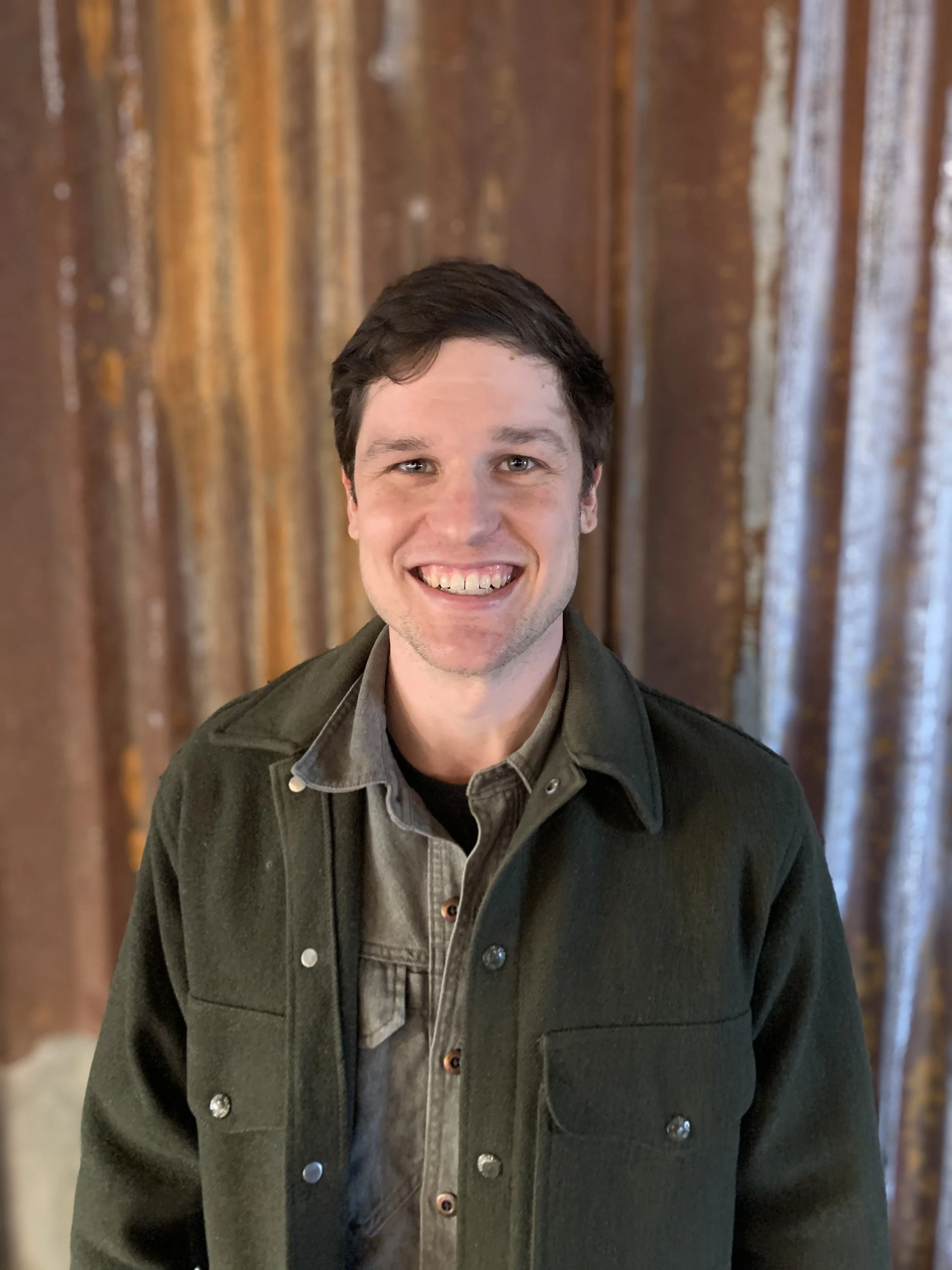 Person smiling in front of a rusty metal wall, wearing a dark jacket and shirt.