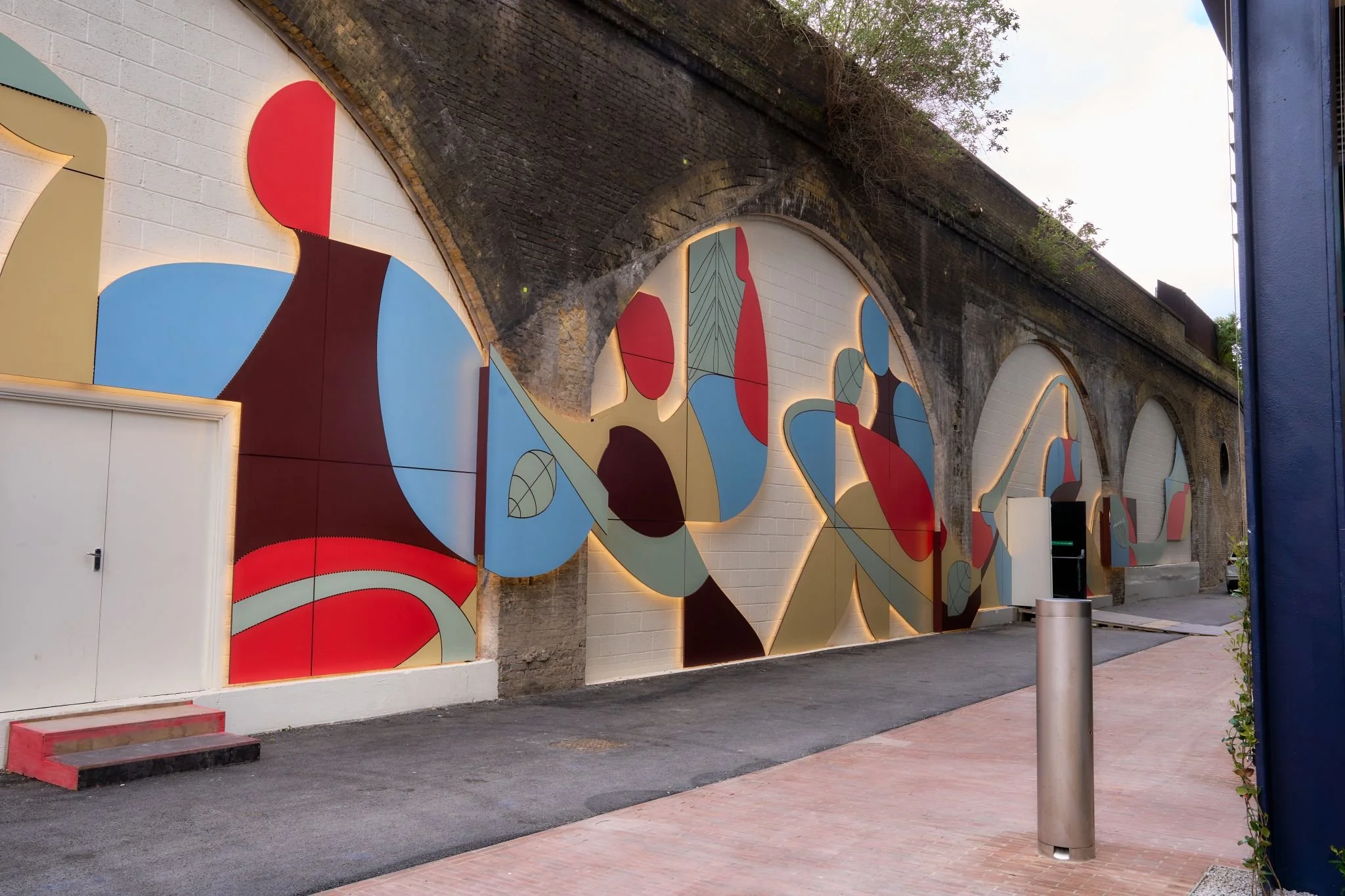Colourful mural installed across a series of railway arches, composed of abstract red, blue, green and cream shapes outlined by soft lighting. The artwork stretches along a pedestrian route between brick walls.