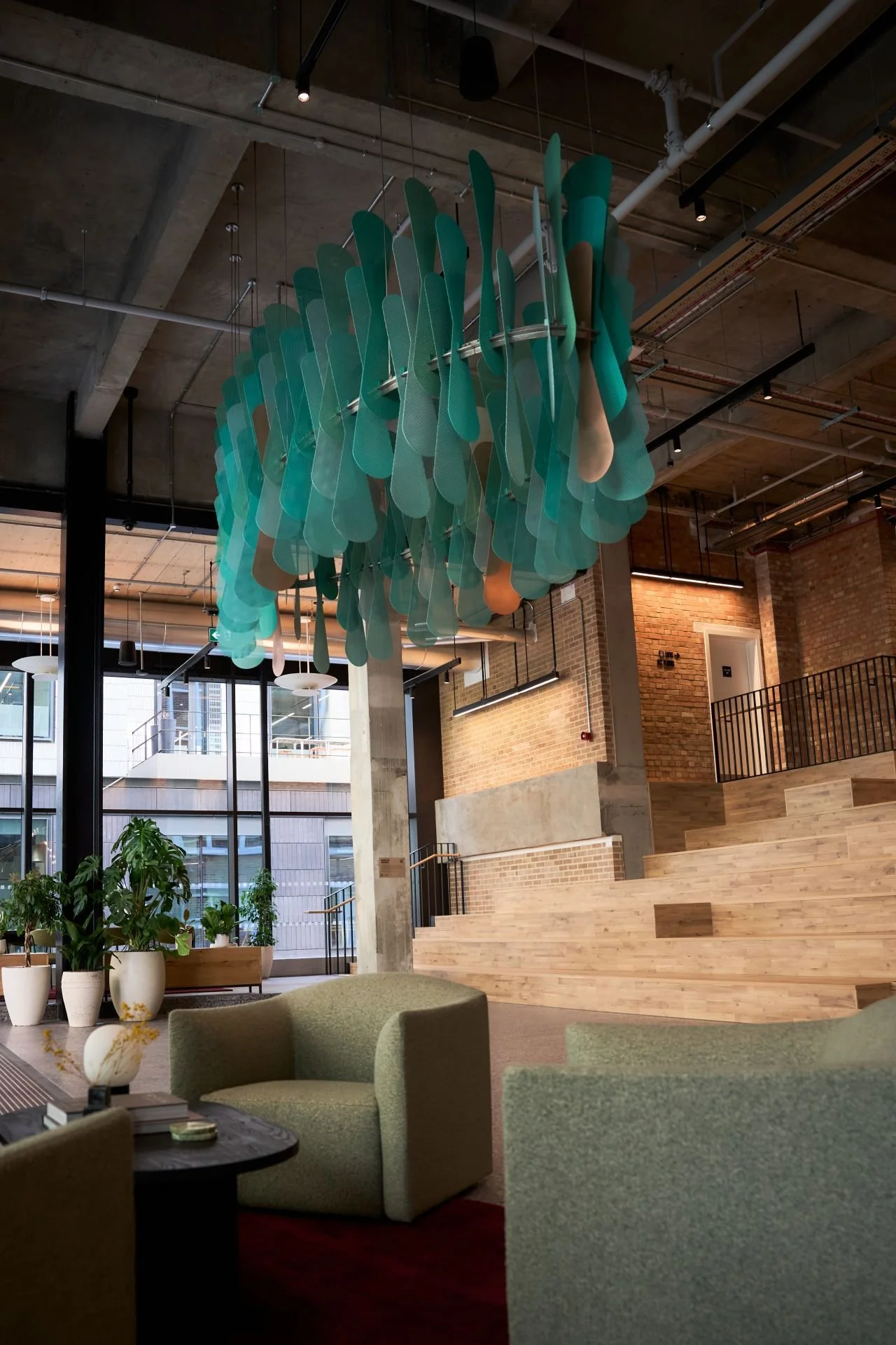 Large suspended artwork made of layered translucent green panels hanging from the ceiling of a spacious timber-and-brick atrium. Seating, plants and stepped wooden terraces are visible below.