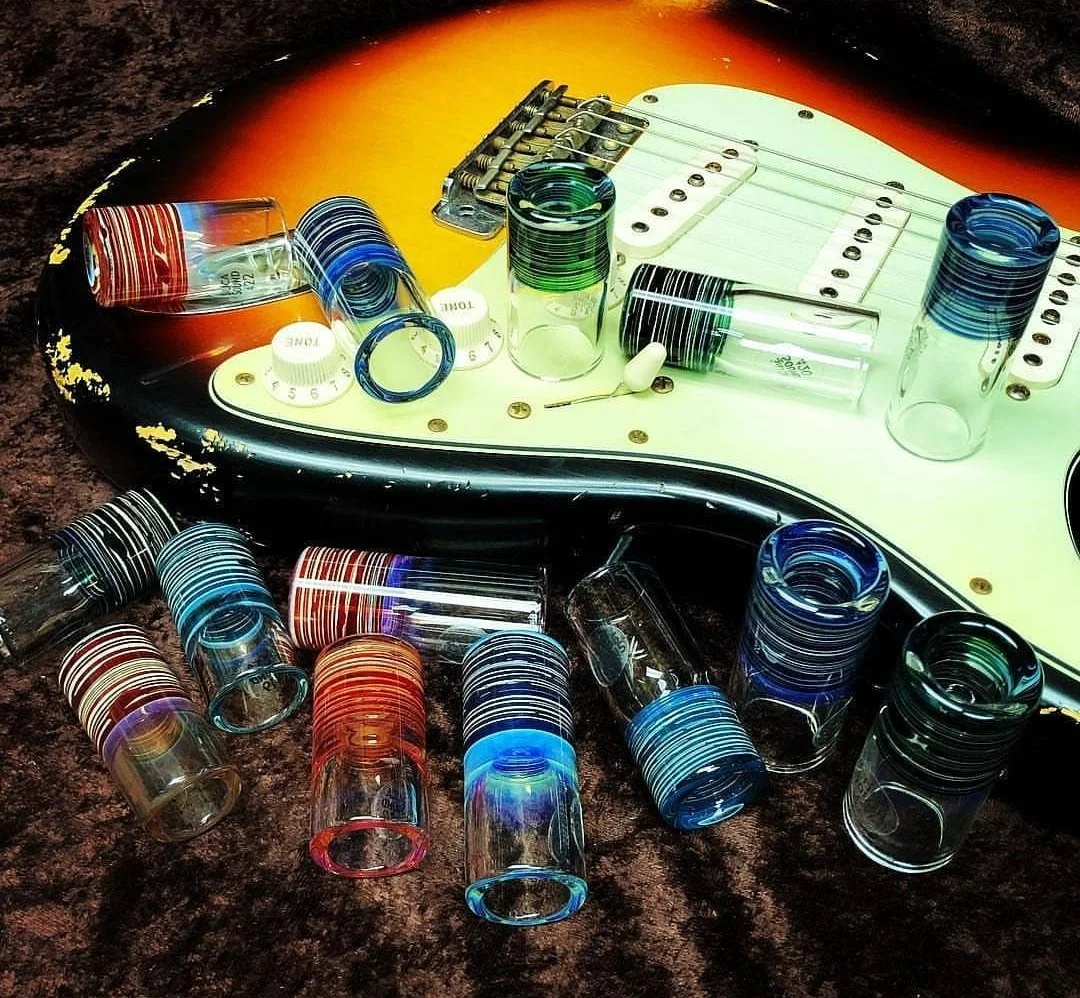 Assortment of handmade glass guitar slides displayed on a Stratocaster-style electric guitar body.