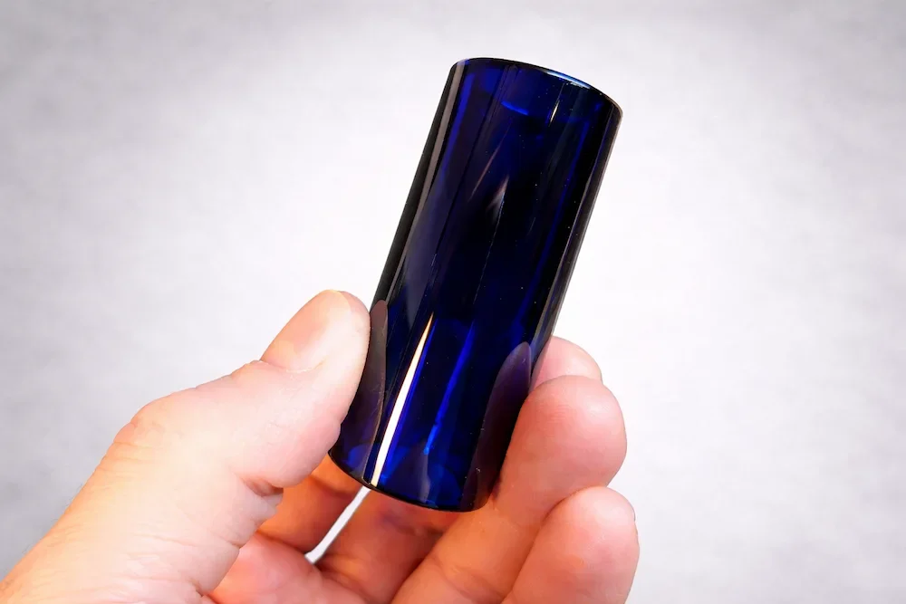 Hand holding a cobalt blue glass guitar slide against a clean light background.