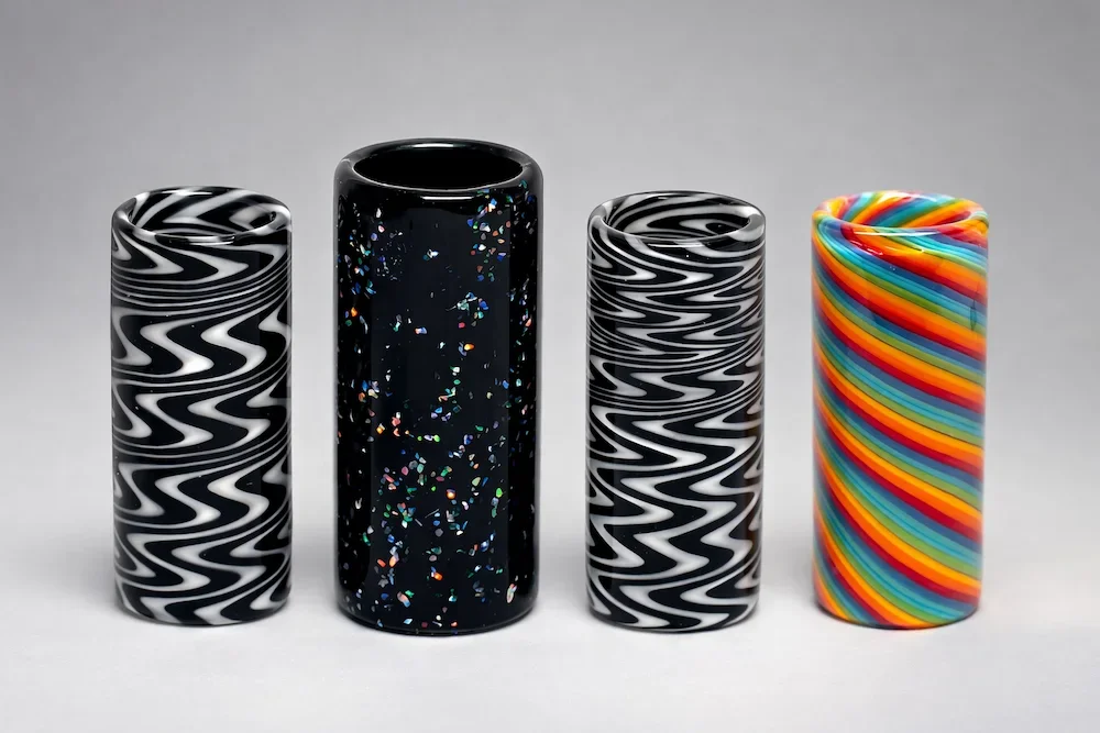 Assortment of one-of-a-kind handcrafted guitar slides with black and white patterns, speckled black finish, and rainbow striped design on a soft gray background.