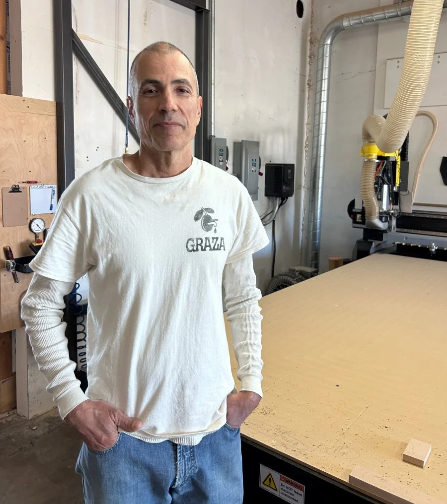 A man with his hands in his jeans pockets wears a white shirt and stands in front of a shop cutting machine