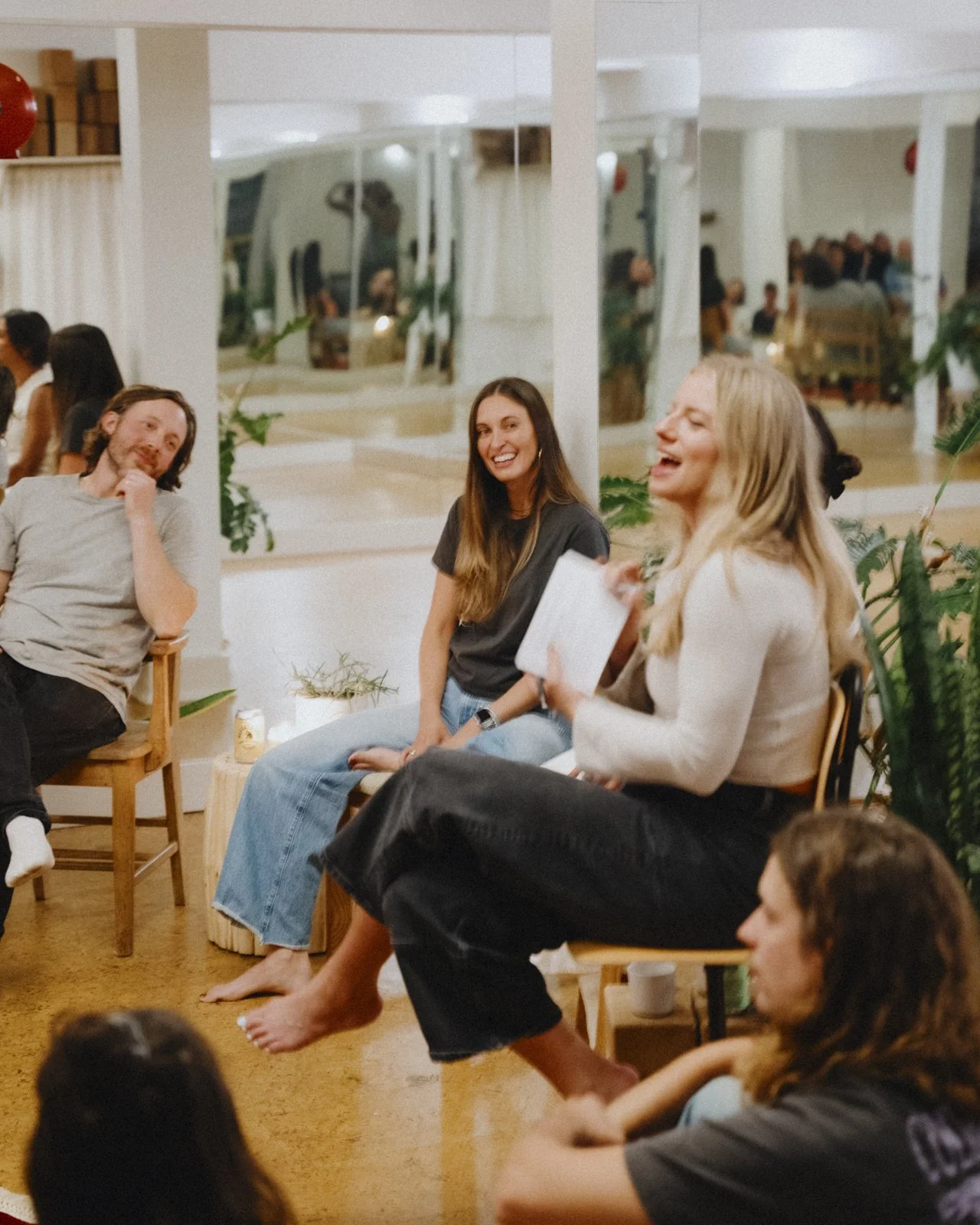 Jess Janz facilitates a panel of creatives in Victoria, BC. They are sitting and laughing together in a cozy indoor space, some on chairs and others on the floor, with plants and large mirrors in the background.