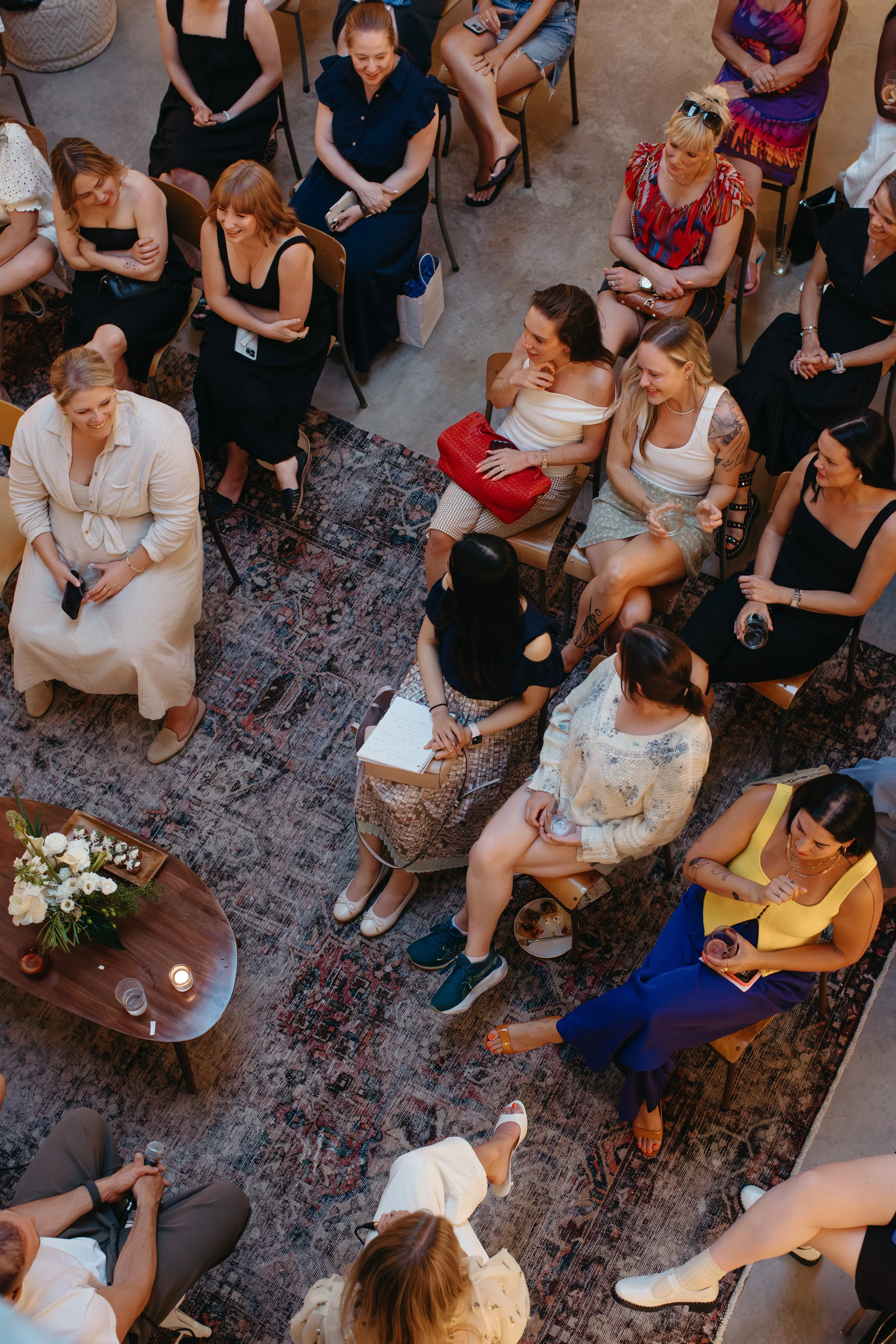 Group of women seated on chairs and sofas at a social gathering or event, some holding drinks, with a decorated table and lit candles in the foreground.