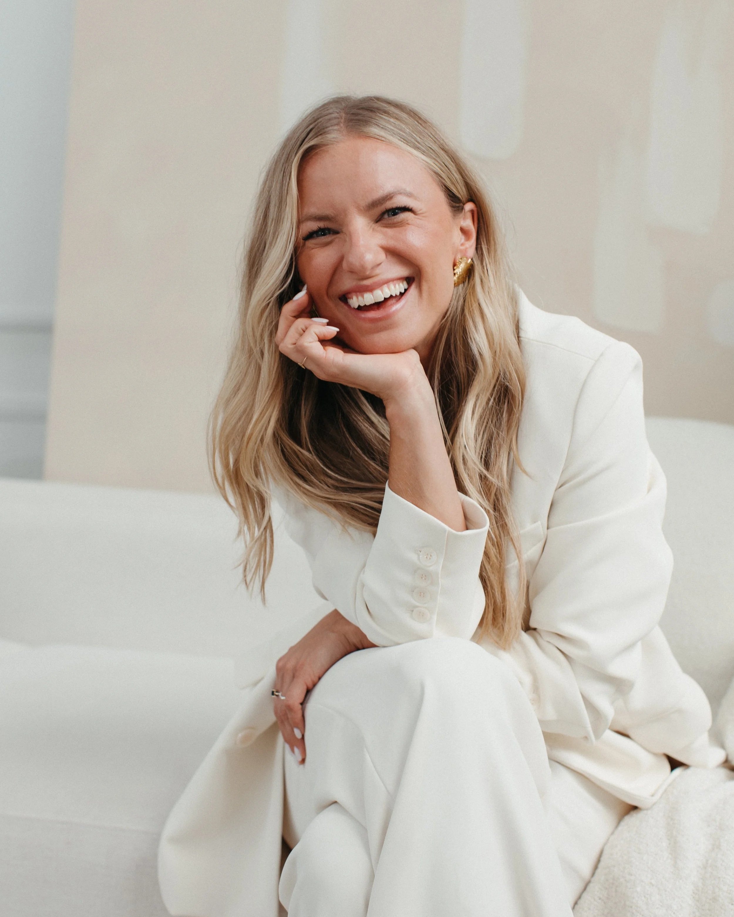 A woman with long blonde hair, wearing a white blazer and gold earrings, is smiling and sitting on a white couch.