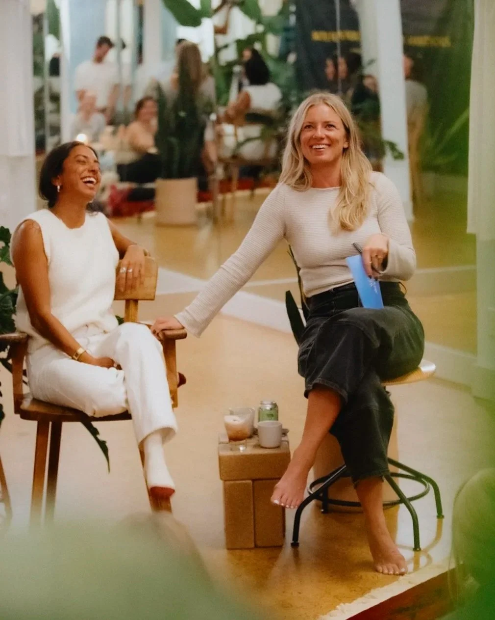 are sitting and laughing together in a social setting. One woman has short dark hair and is wearing white clothing, while the other has long blonde hair and is wearing a light-colored sweater and dark jeans. They appear to be enjoying a friendly conversation, with a small table in front of th