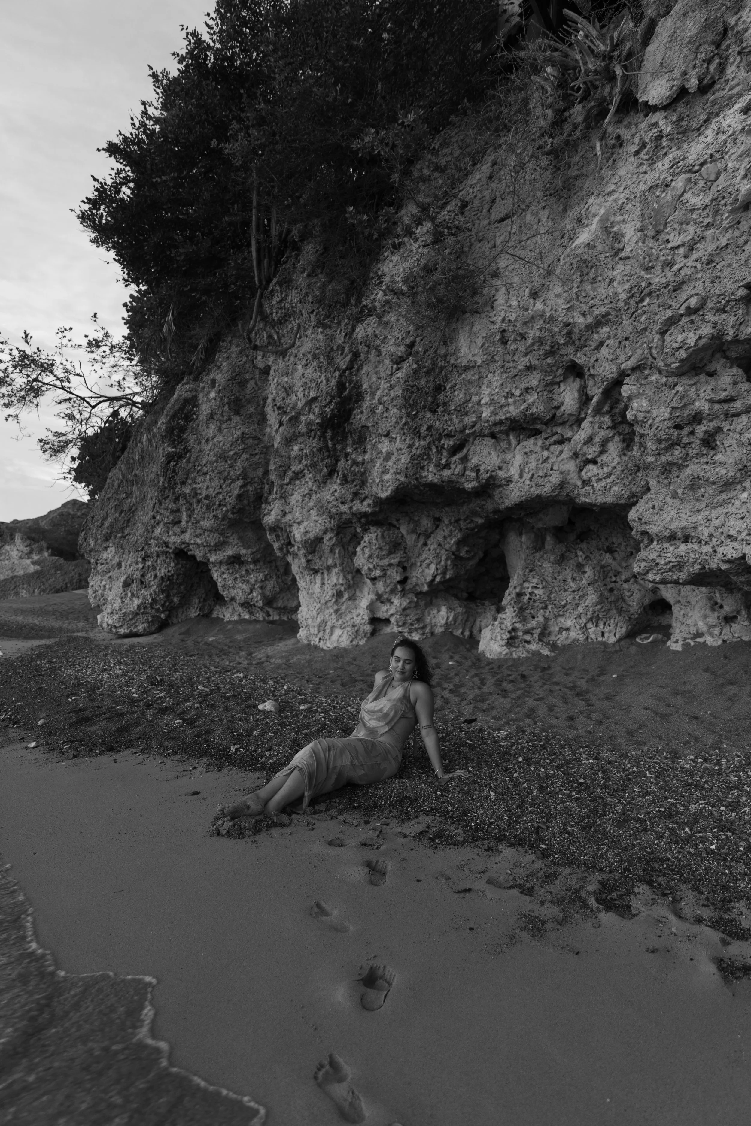 A woman lounging on a sandy beach, sitting against rocks with greenery at the top, near the water's edge, with footprints on the sand in front.