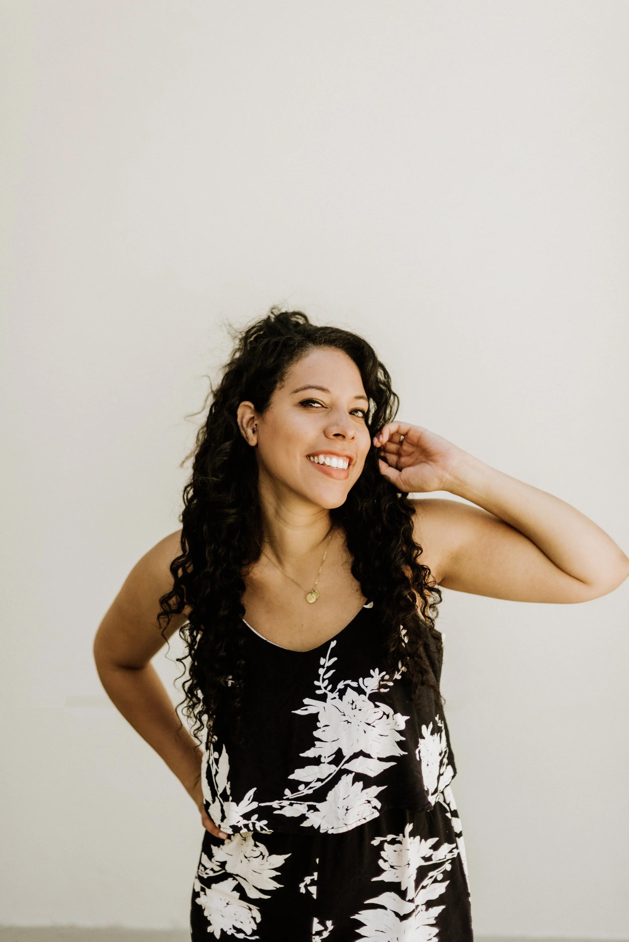 woman with dark curly hair in navy blue jumper looking directly at the camera with a white wall background