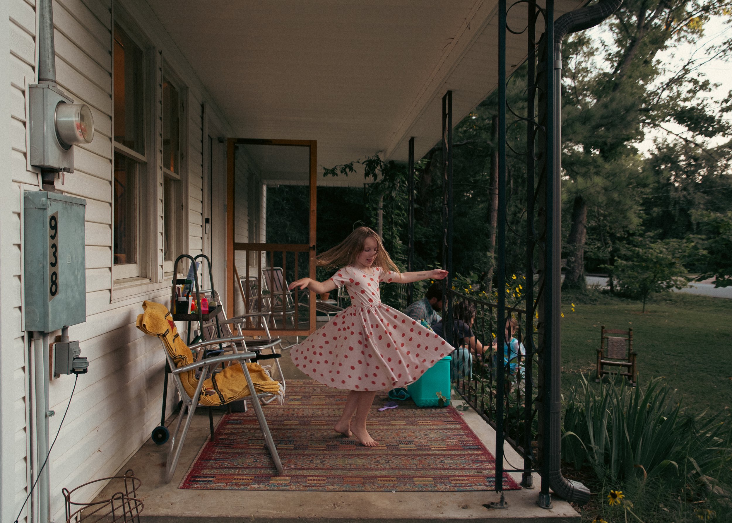 A young girl in a white dress with red polka dots twirling on a porch with a colorful rug. There are chairs, a yellow towel, and household items around her. She appears to be dancing happily, with outdoor greenery and trees visible in the background.
