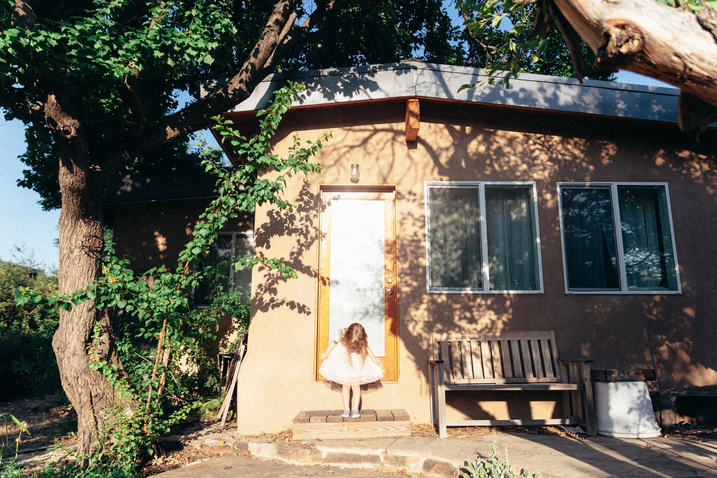 A young girl in a white dress stands on a small wooden porch, facing a door with a white board covering it, in front of a beige house with large windows. Shadowed trees are visible, casting shadows on the house exterior.