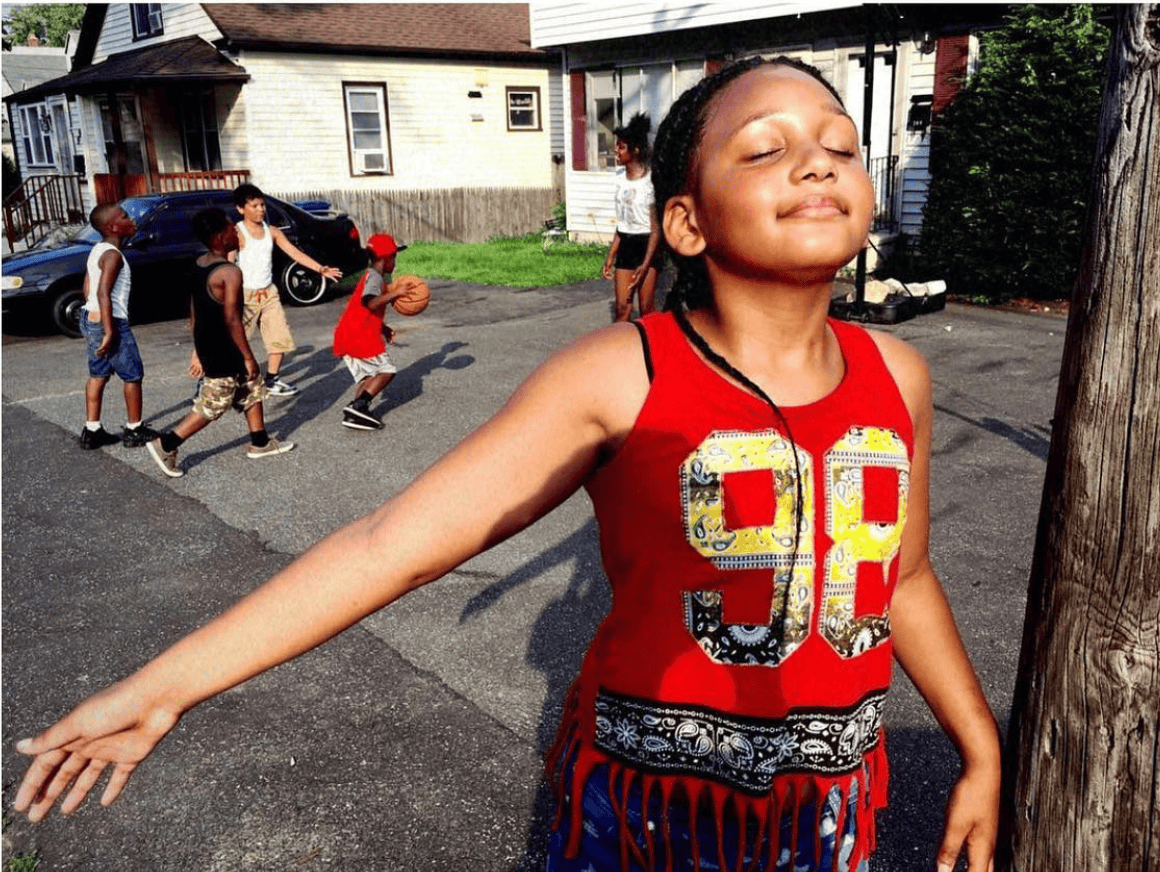 A young girl in a red tank top with the number 99 on it stands with her eyes closed and arms outstretched, smiling, in front of a group of children playing basketball on a driveway. The background features houses, parked cars, and trees.