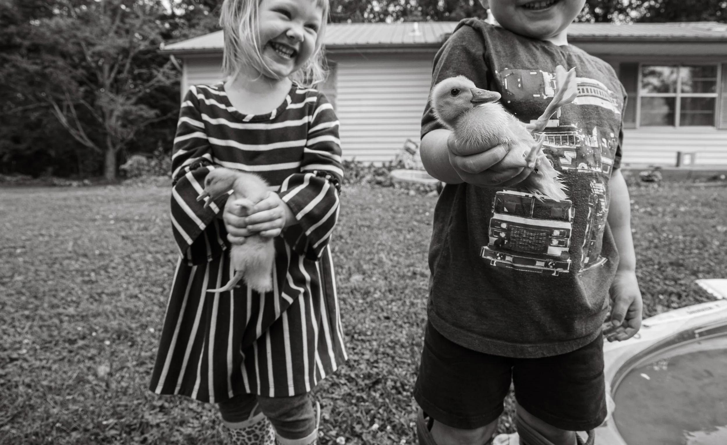 Two children smiling and holding ducklings outdoors in front of a house.