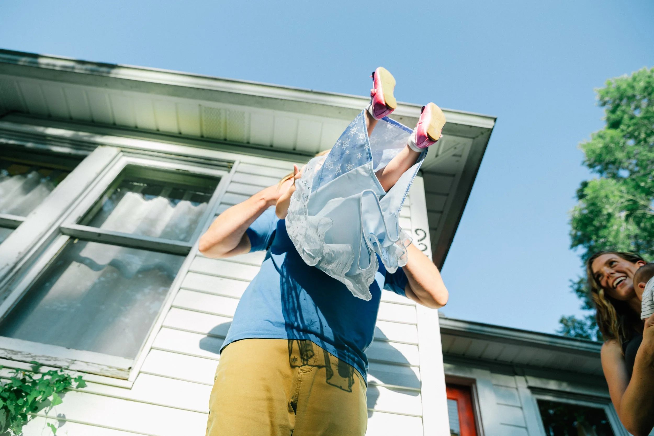 Person holding a baby and a woman smiling outside a house on a sunny day.