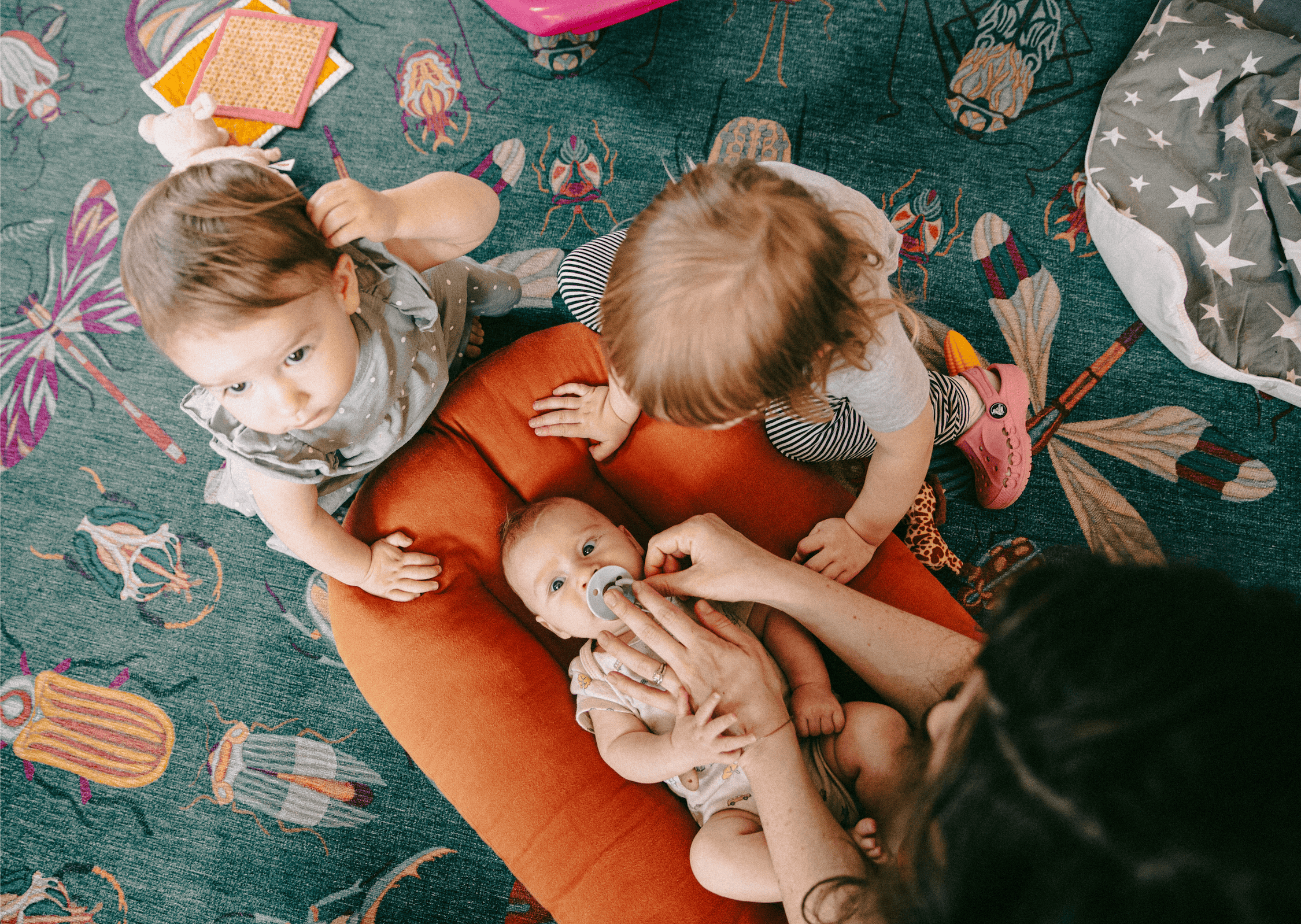 View from above of three young children and an adult, with the adult helping a baby with a pacifier, on a colorful carpet with bugs, dragonflies, and insects pattern.