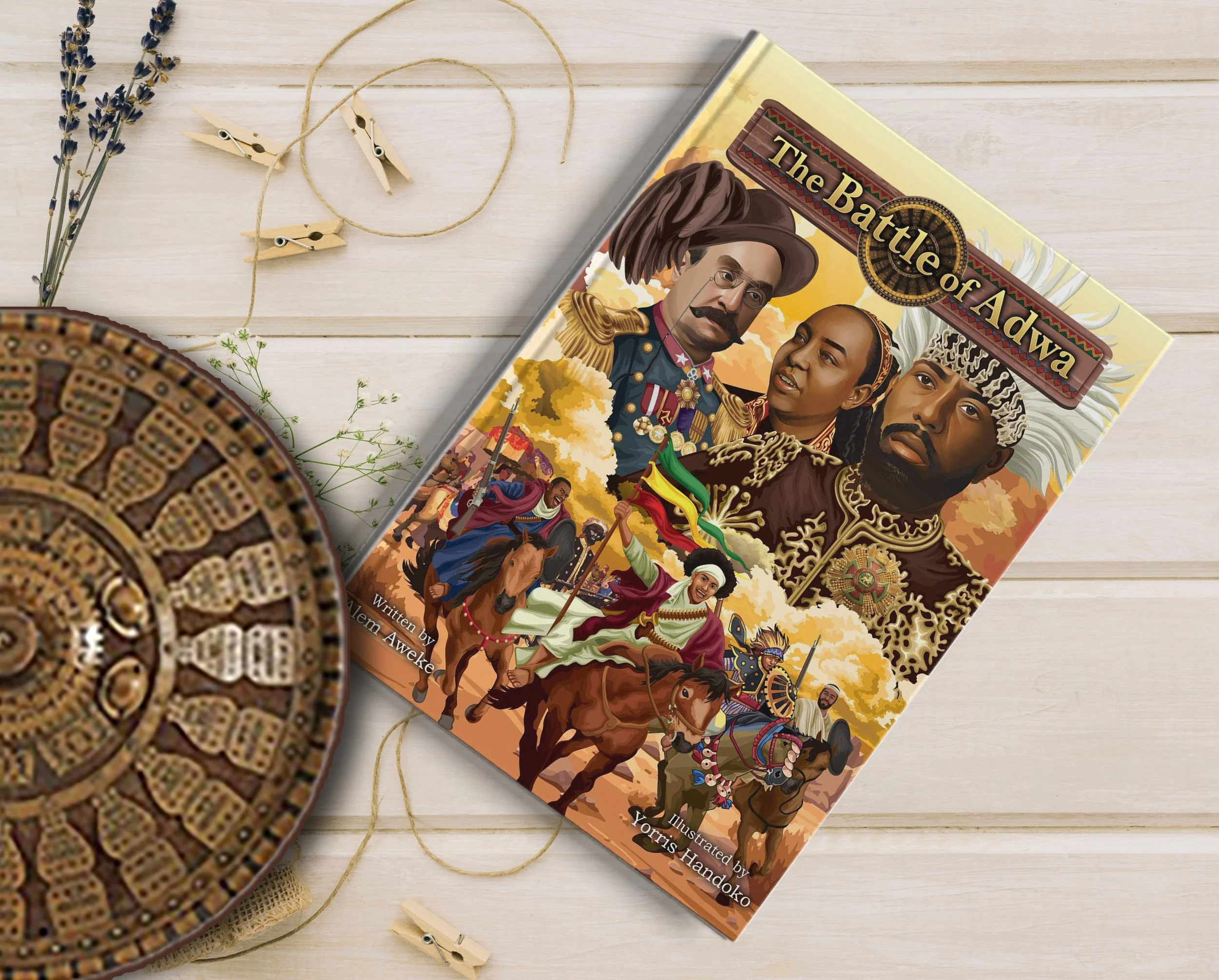 Book cover titled "The Battle of Adwa" by Aem Awelke featuring historical African figures in traditional attire on a yellow background, placed on a light wooden surface with decorative items around.