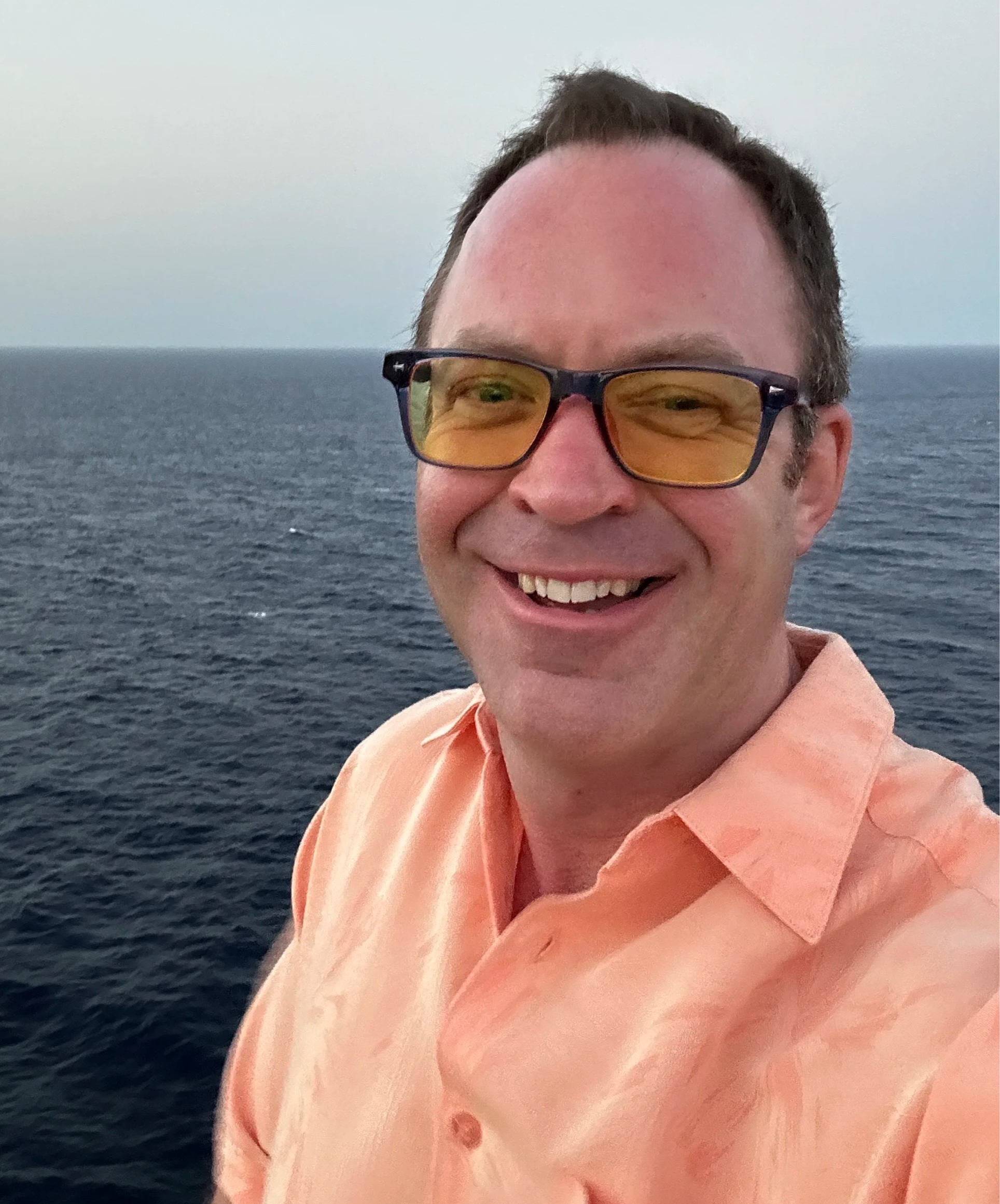 A man smiling outdoors near the ocean, wearing glasses with orange lenses and a peach-colored shirt.