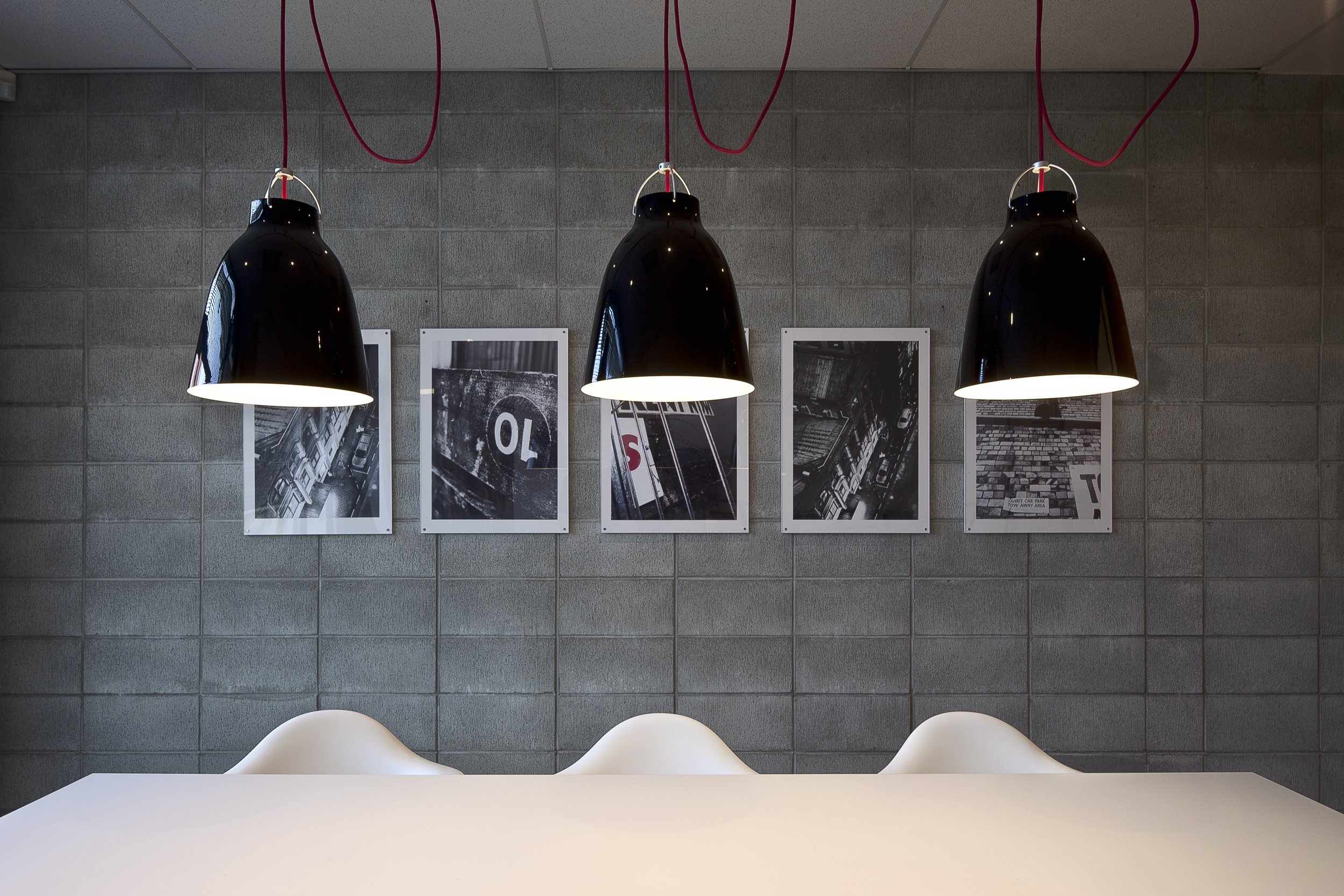 Modern dining area with three black pendant lights hanging over a white table and chairs. The gray tiled wall in the background features five abstract black and white photographs.