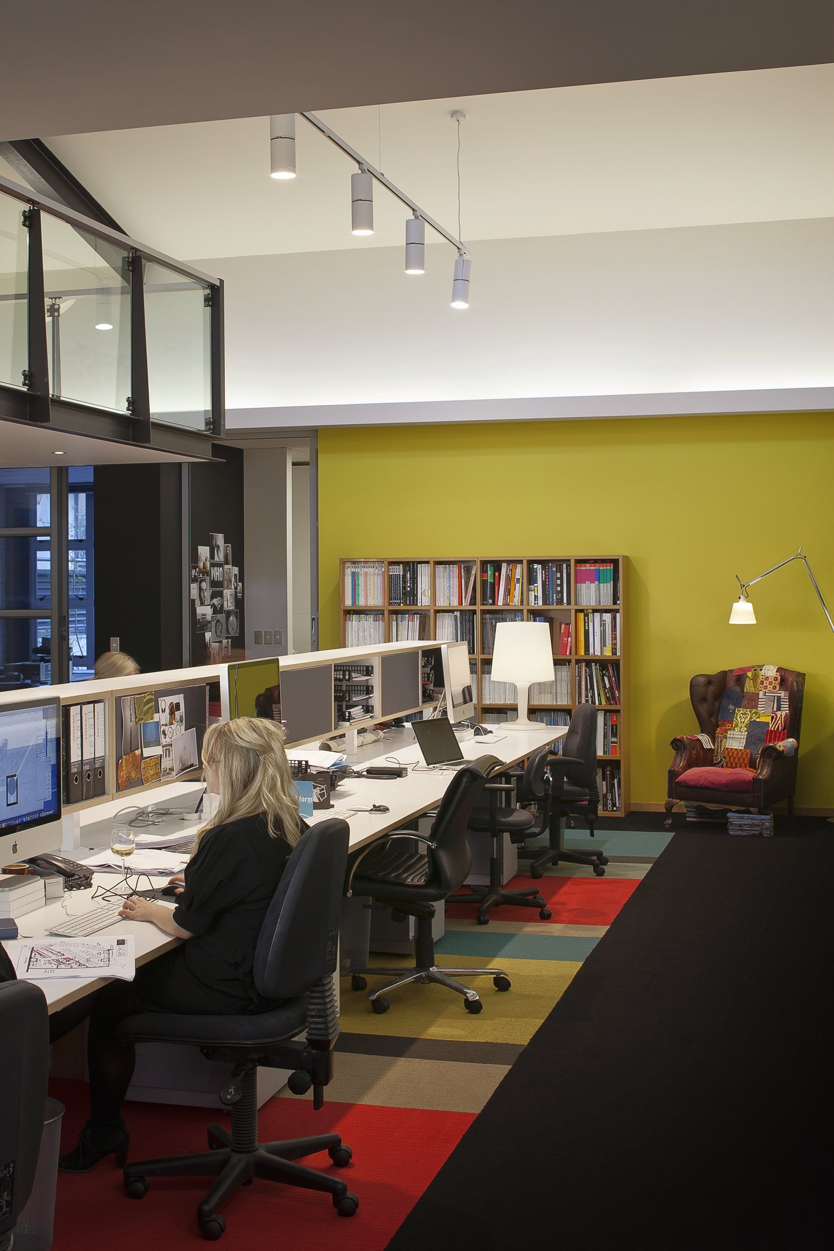 Modern office space with a row of desks and computers, colorful carpet, shelf with books, bright green wall, and ceiling track lights.