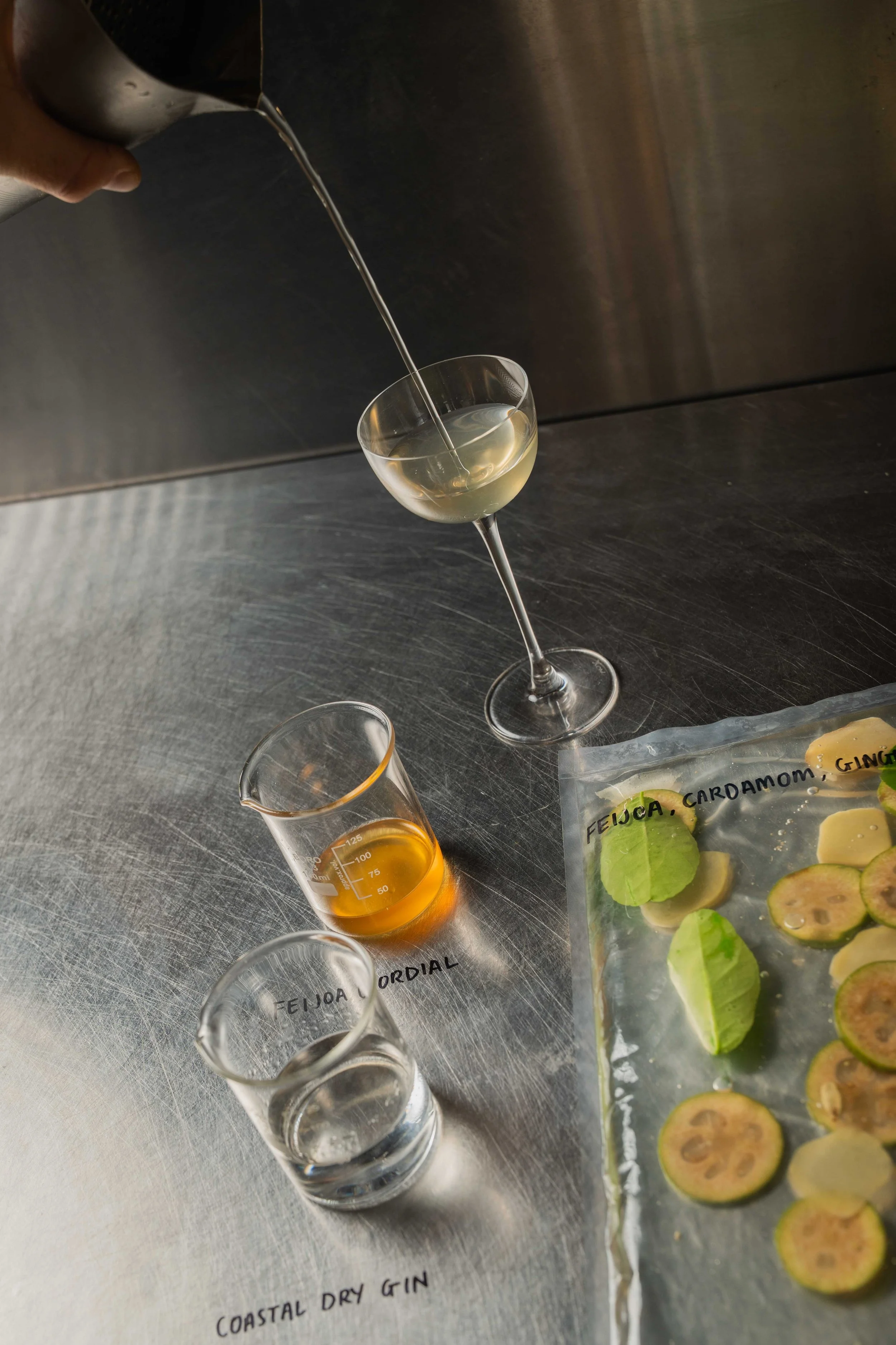 A person pouring a clear liquid into a cocktail glass on a metal surface. There is a small beaker with orange liquid and sliced limes on the table, along with a plastic bag labeled with ingredients such as feijoa, cardamom, and ginger.