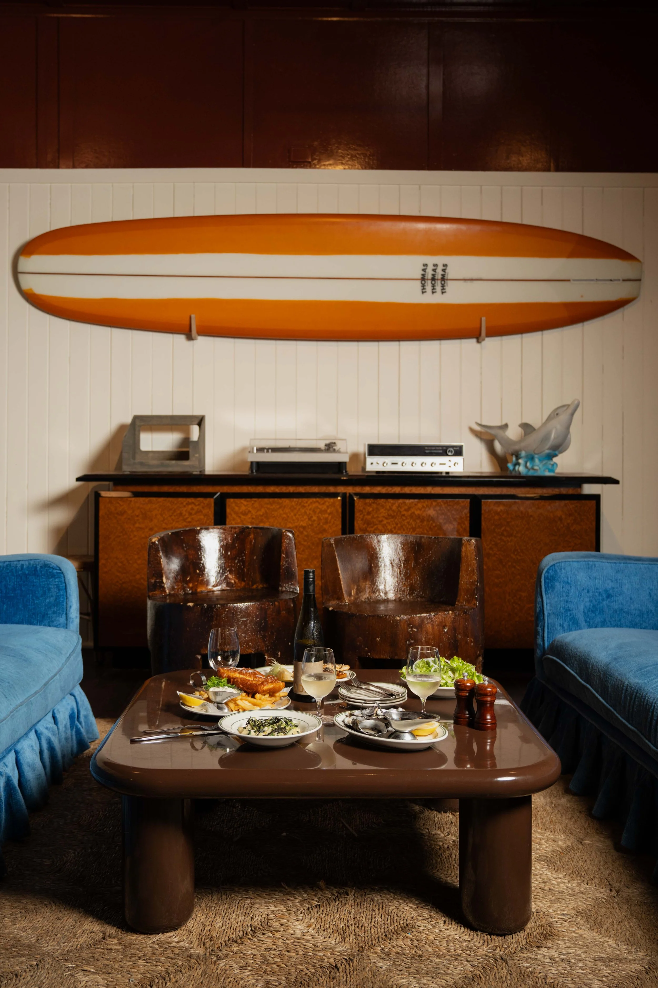 A dining room scene with a brown table set with plates of food and drinks, surrounded by blue velvet and brown wooden chairs. In the background, a wall-mounted surfboard with the name 'Thomas' written multiple times is visible, along with decorative 