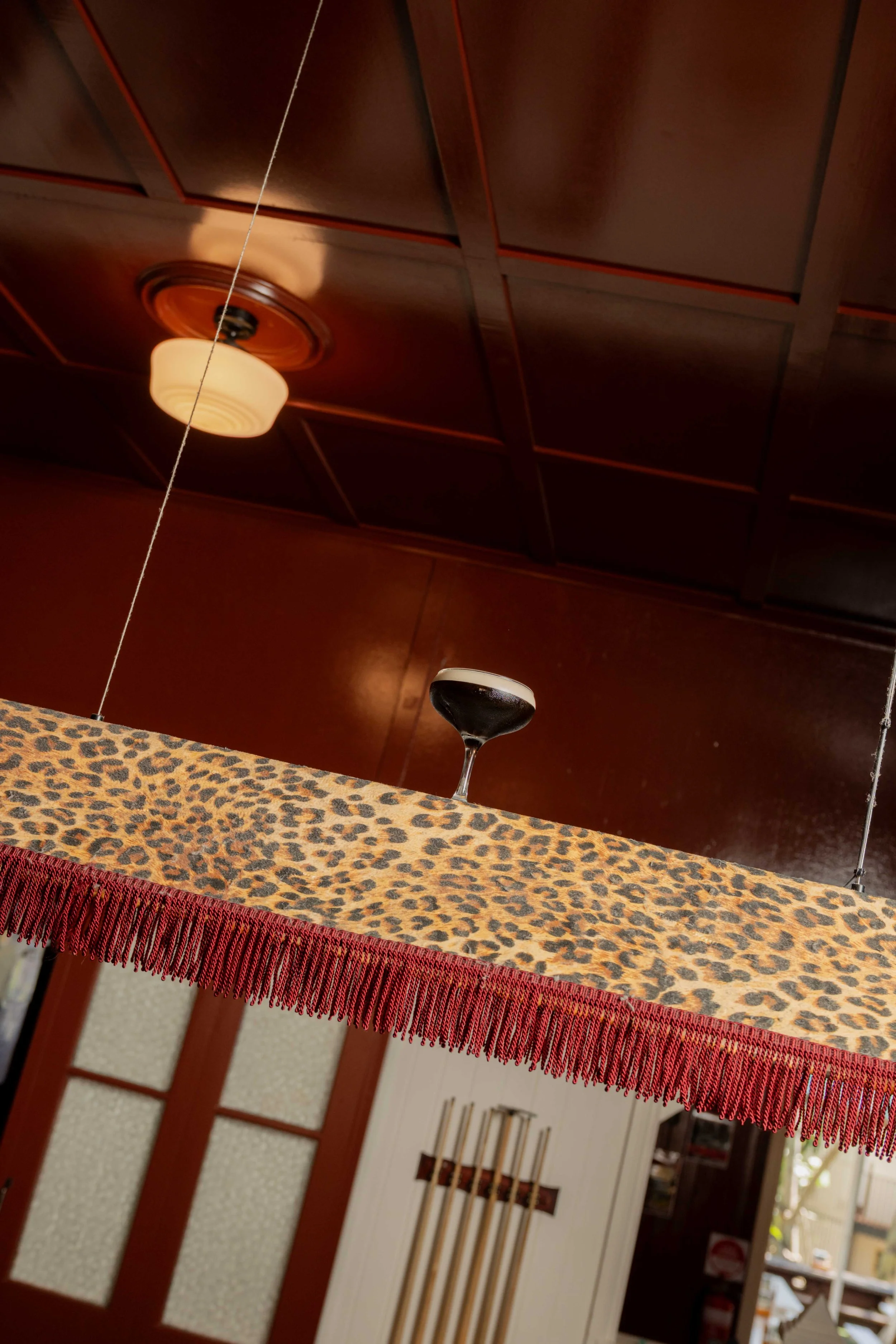 Close-up of a decorative table with a leopard print tablecloth and a red fringe trim, in front of a wooden cabinet with a cross design on the door, inside a room with wood paneling and a light fixture on the ceiling.