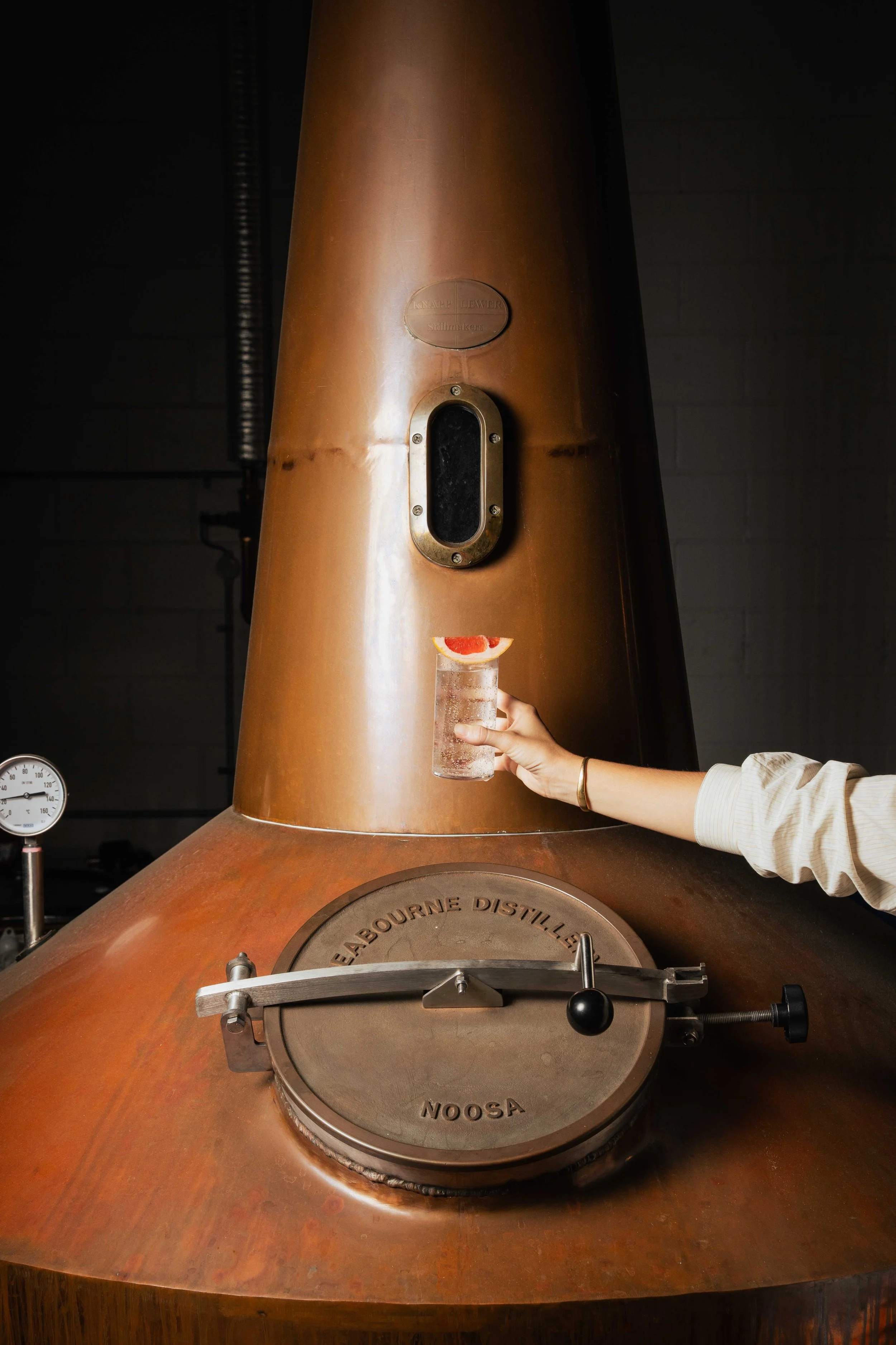 A copper still in a distillery with a hand holding a glass of clear spirit garnished with a slice of grapefruit in front of it.