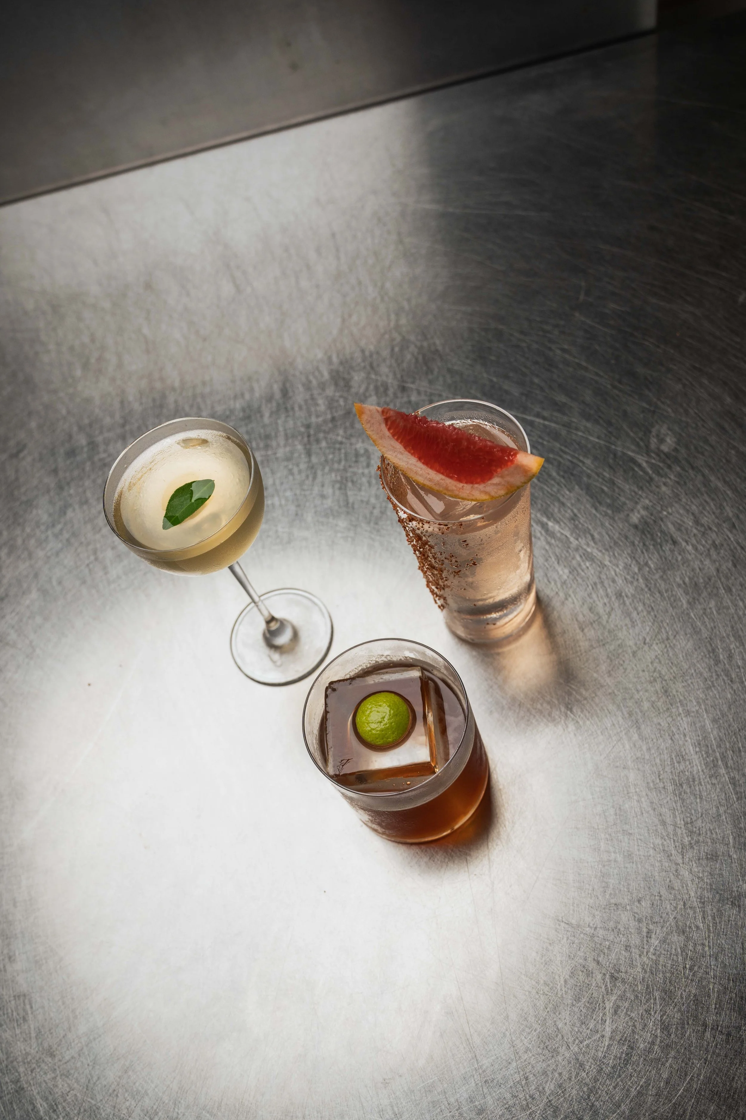 Three cocktails on a metallic bar counter: one with a lime wedge, one with a grapefruit slice and sugar rim, and another with a leaf garnish.