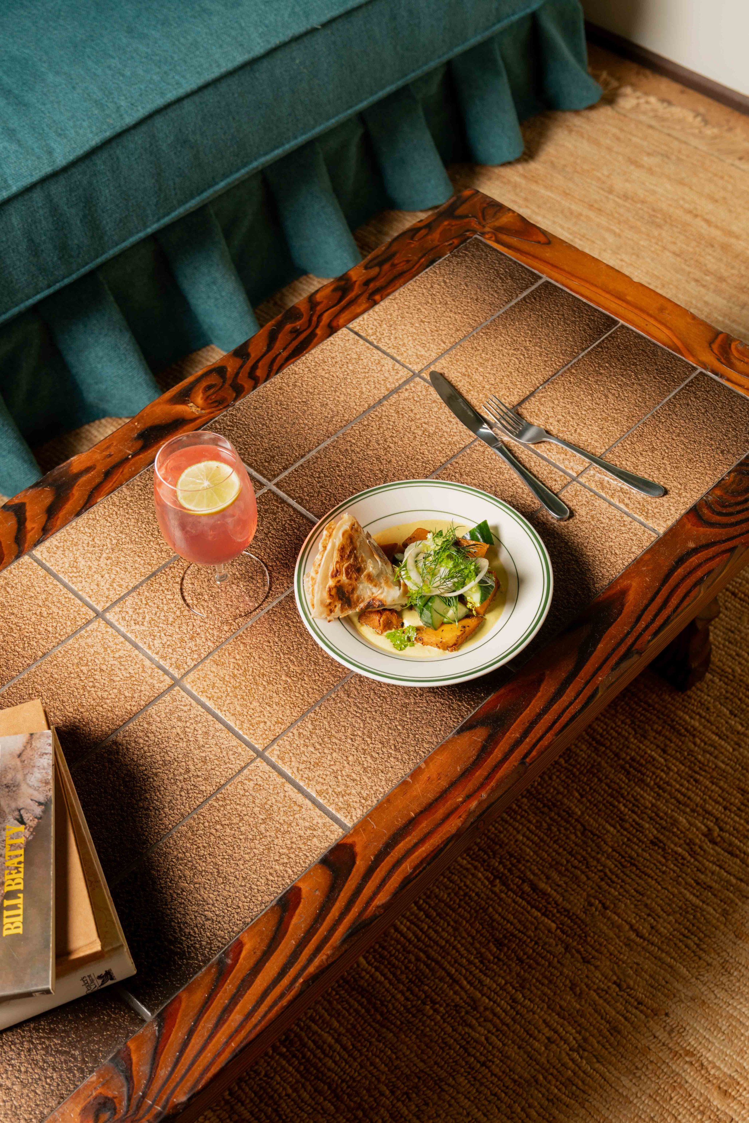 A dining table with a tiled surface, holding a plate of salad and a slice of pie, a glass of lemon water, a fork and knife, and a wooden edge, with a green upholstered bench in the background.