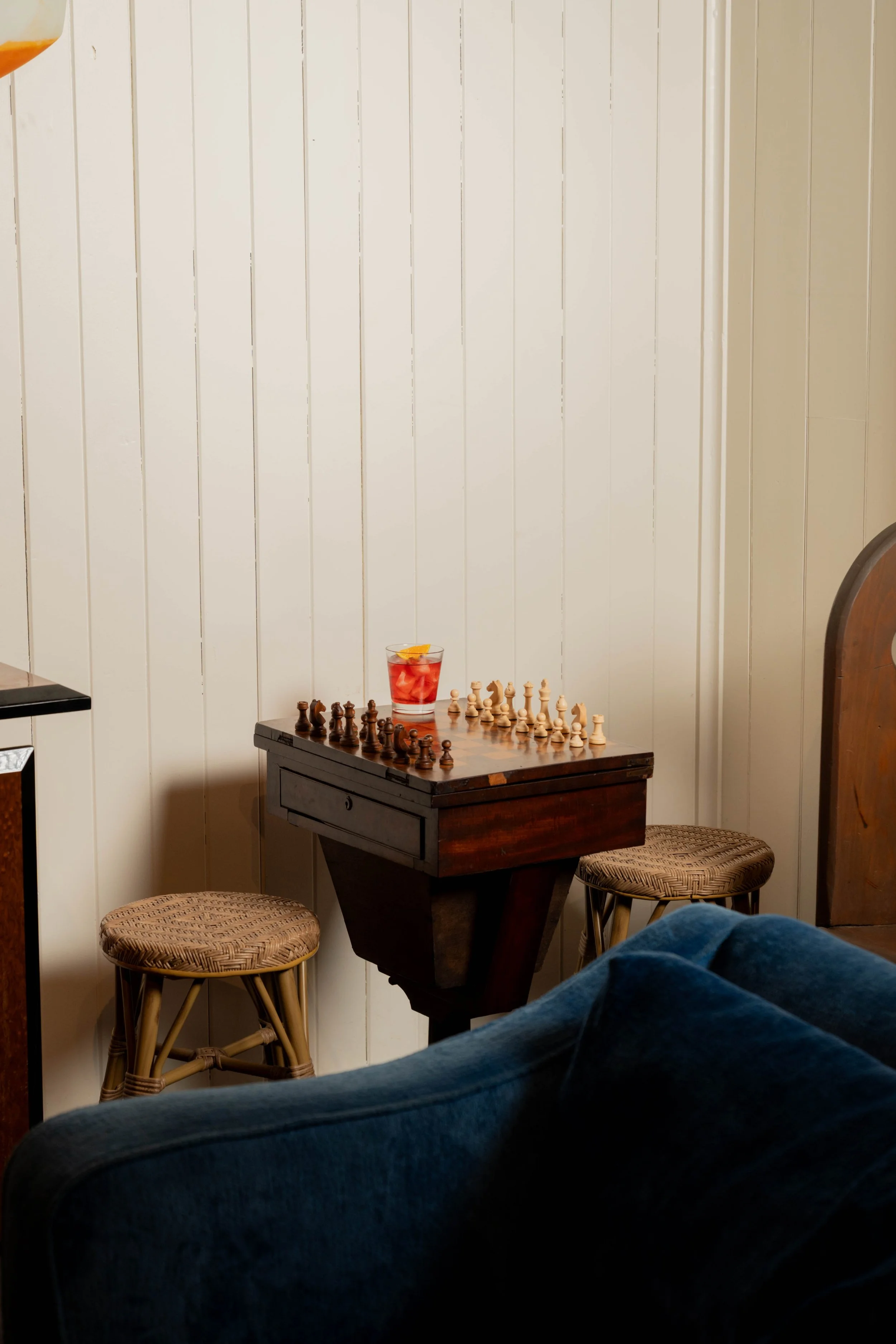 A small wooden table with a chess set and a glass of red beverage with a slice of orange on top, two woven stools, and a white paneled wall in the background.