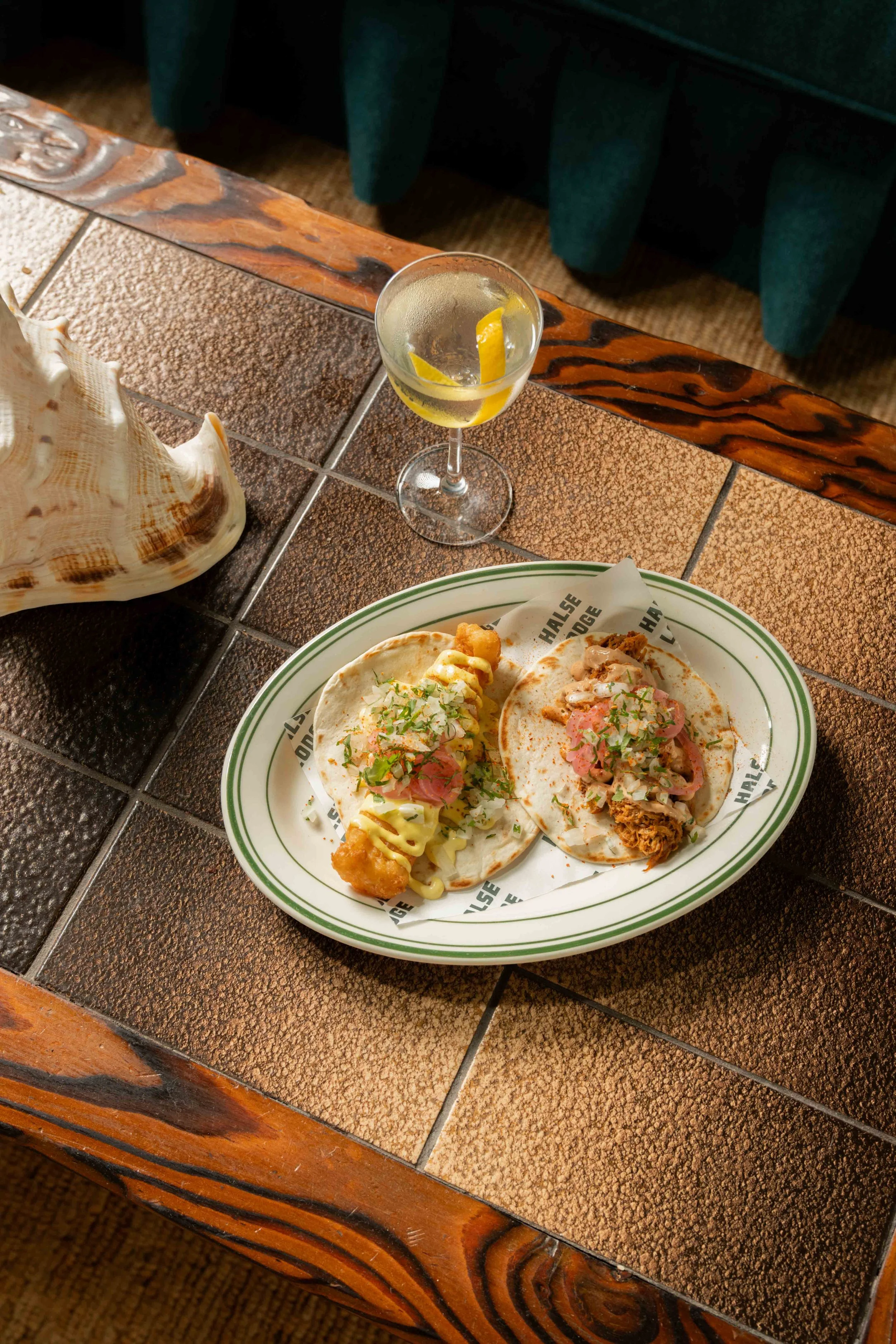 A plate with two tacos topped with chopped onions, cilantro, and sauce, placed on a rustic wooden table with tile insets, alongside a drink garnished with lemon wedges.