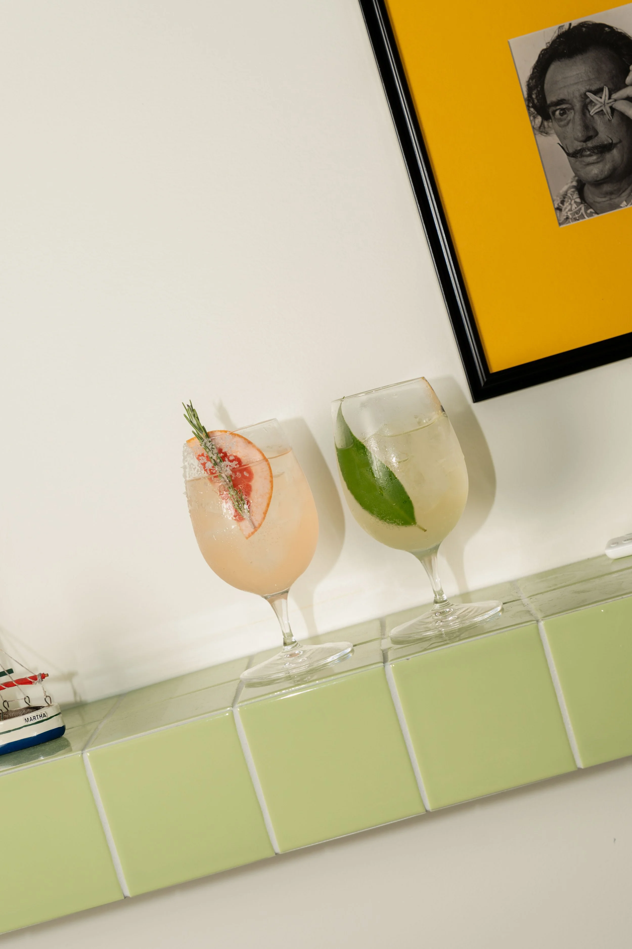 Two cocktails on a counter with a framed black-and-white photo on the wall. The cocktail on the left has a grapefruit slice and rosemary garnish, and the one on the right has a lime wedge.