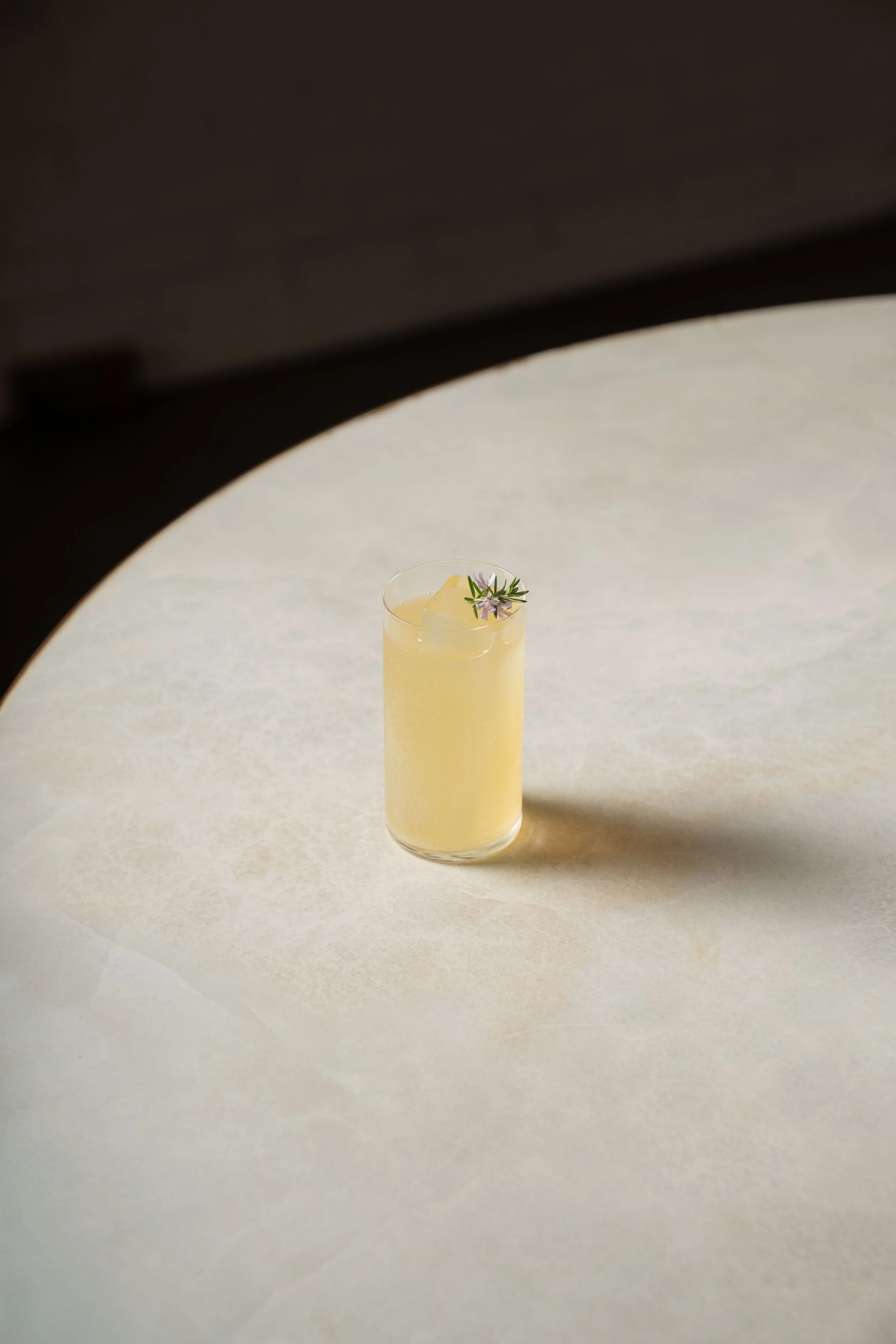 A tall glass filled with a pale yellow beverage, garnished with a sprig of purple flowers and green leaves, on a smooth, light-colored surface with a dark background.