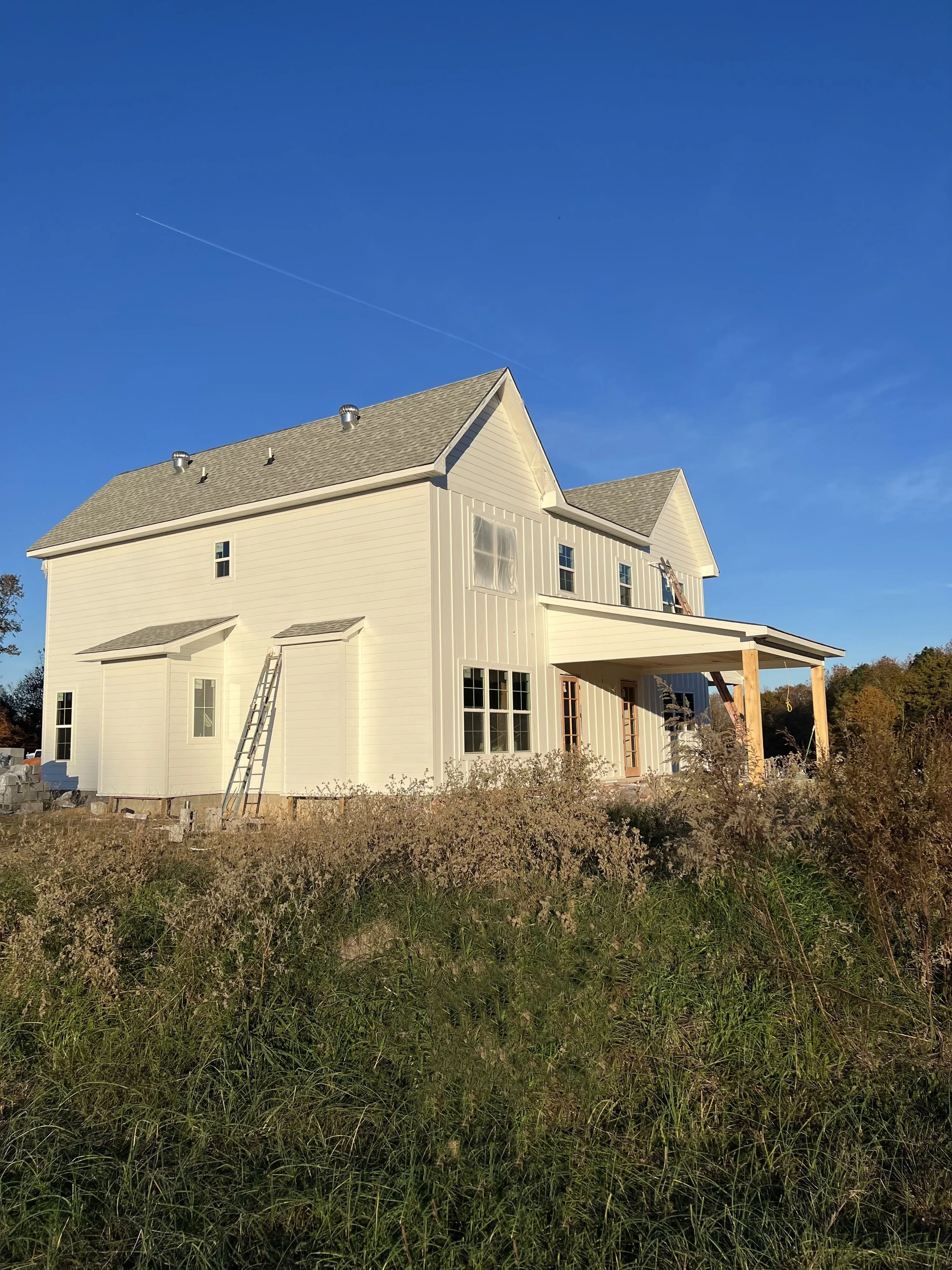 A white two-story house under construction with a front porch and multiple windows, surrounded by tall grass and bushes, against a clear blue sky.