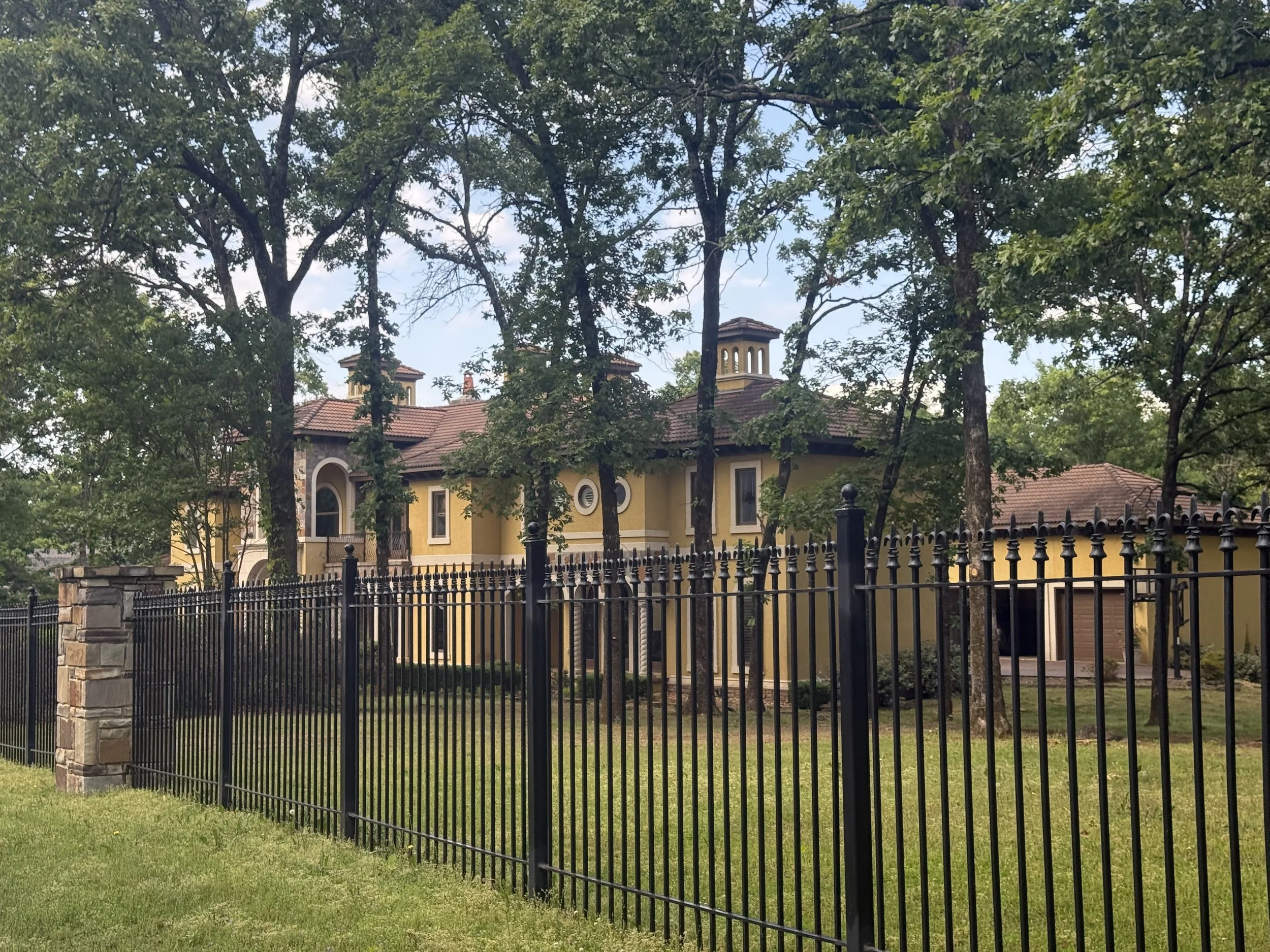 A yellow house with tiled roof and chimneys is seen behind a black metal fence. Tall trees with green leaves surround the house, and the sky is partly cloudy.