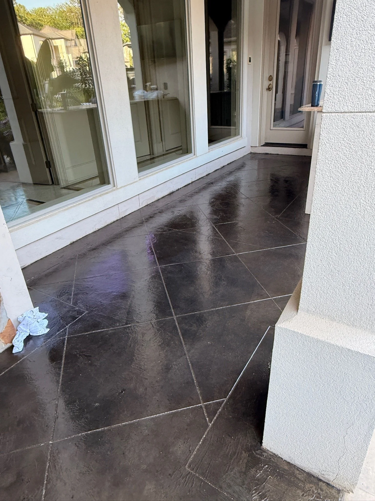 A porch area with dark shiny tiled flooring, beige walls, large glass sliding doors, and a screen door leading inside a house. There is a small ledge on the right side with a dark tumbler glass on it.