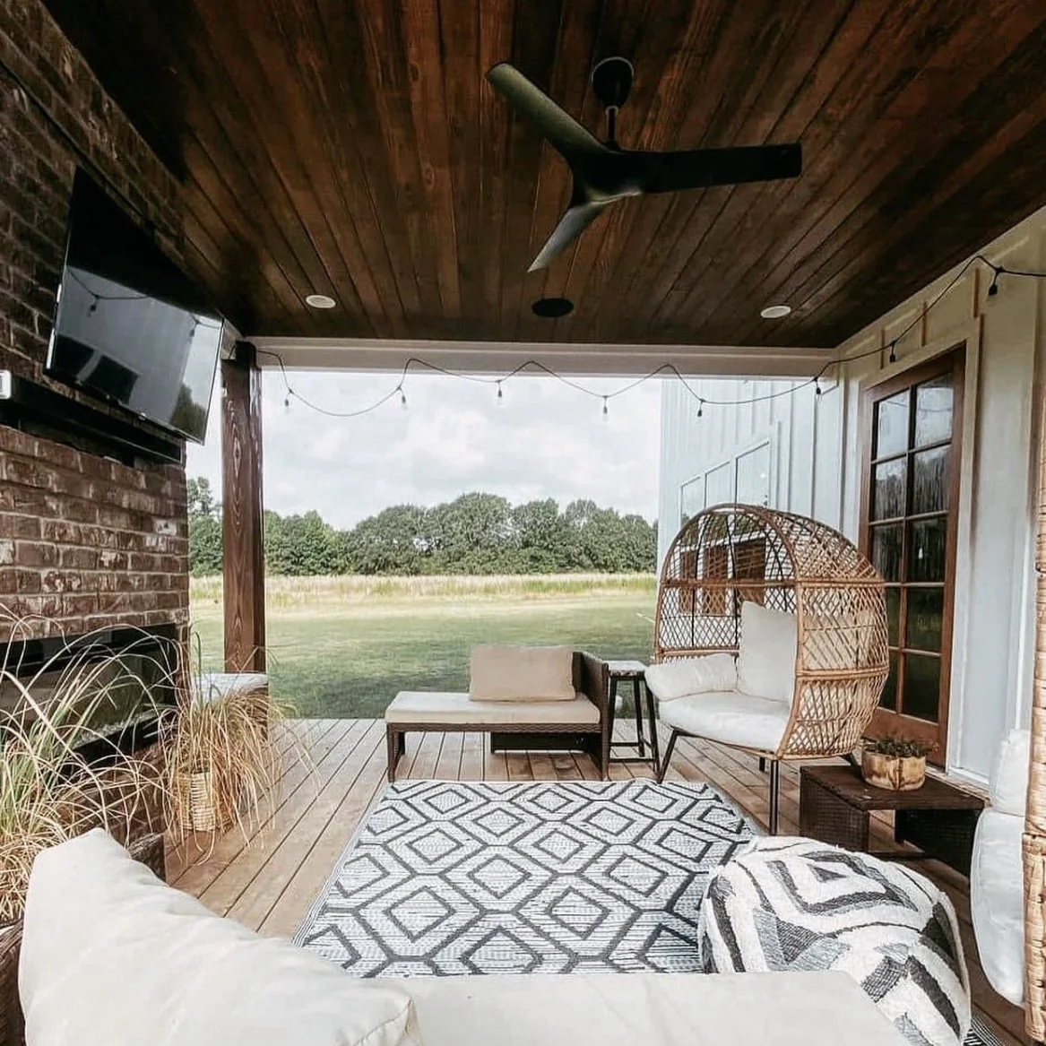 Covered outdoor patio with wooden ceiling and brick wall, ceiling fan, string lights, outdoor seating including a wicker hanging chair with cushion, a sectional sofa, a patterned rug, a flat-screen TV, and a view of a grassy field and trees in the ba