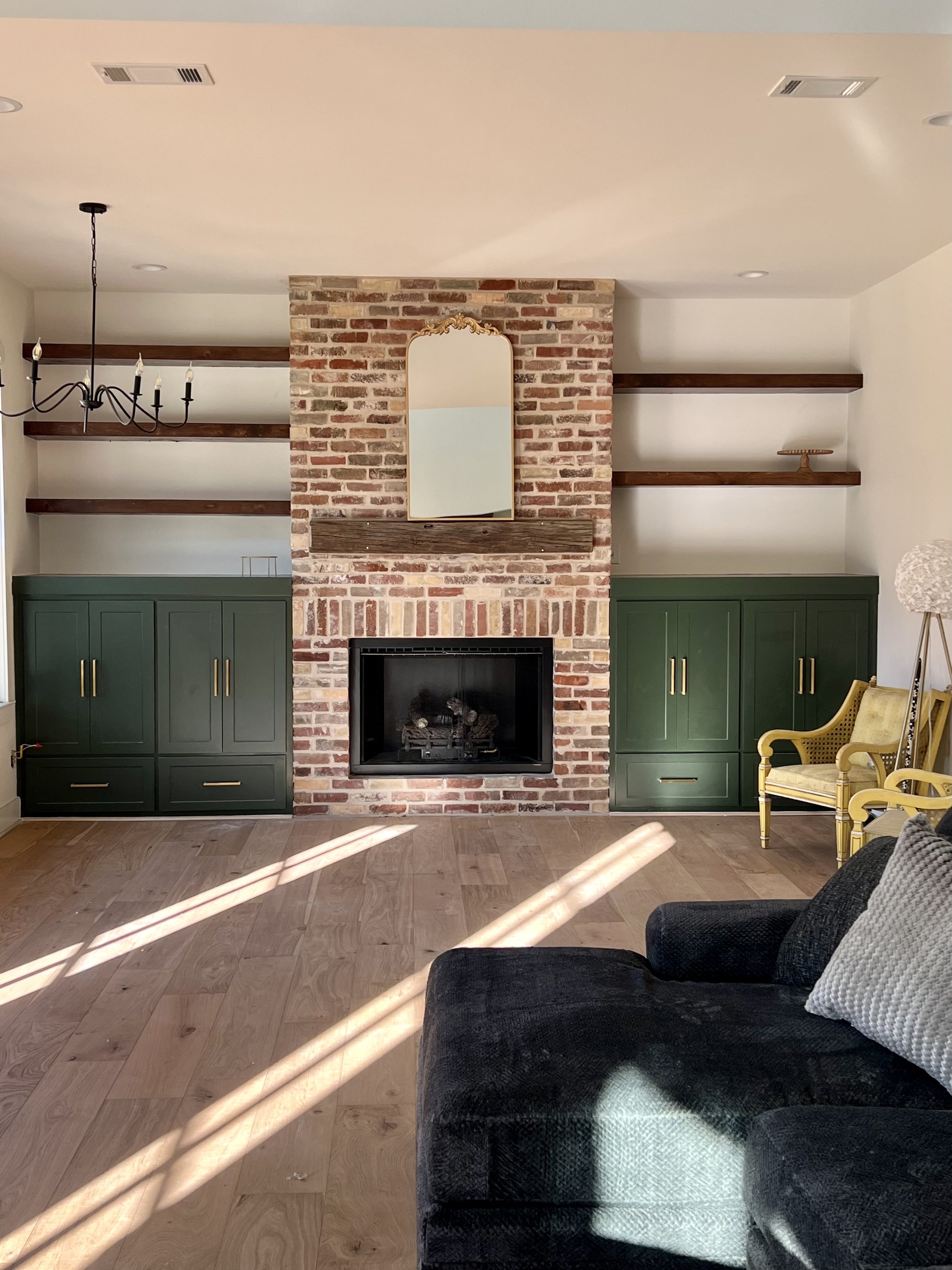 Living room with brick fireplace, gold-framed mirror, dark green cabinets, wooden shelves, black sofa, yellow armchair, and sunlight on wooden floor.