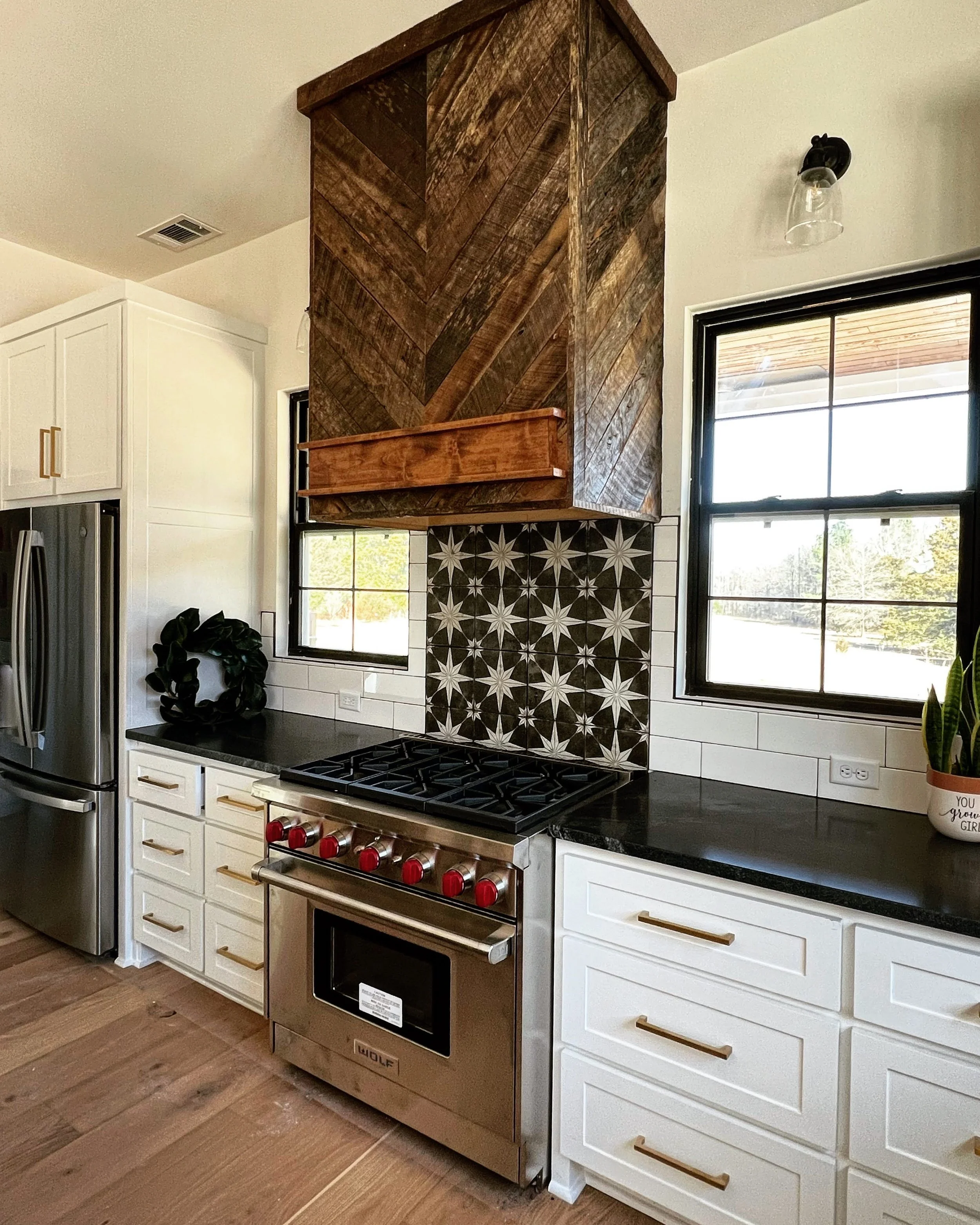 Kitchen with black countertop, white cabinets with gold handles, stainless steel Wolf oven, decorative black and white star-patterned tile backsplash, wood range hood, windows with black frames, plant in white pot, and wooden flooring.