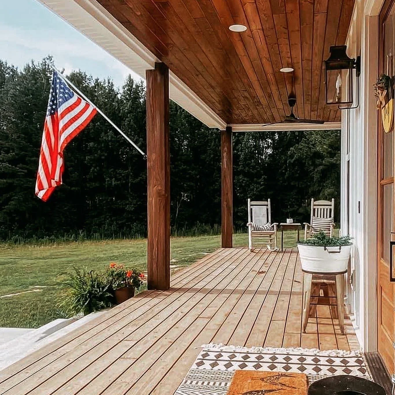 A wooden porch with two chairs and a small table, an American flag is hanging from a pole on the left, and potted plants and flowers are on the porch, with a grassy yard and trees in the background.